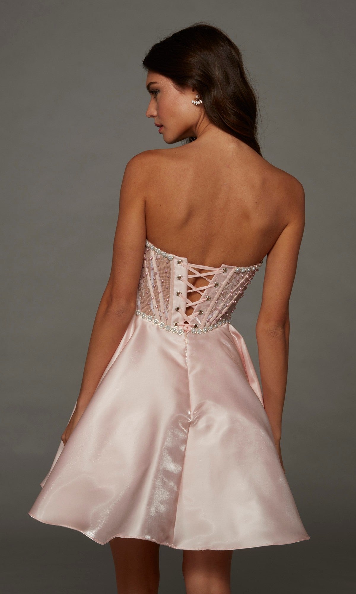 Model wearing the Short Homecoming Dress: Alyce 30088, which features a strapless silhouette with a satin, light pink corset bodice adorned with delicate pearl embellishments and a lace-up back, standing against a neutral background.