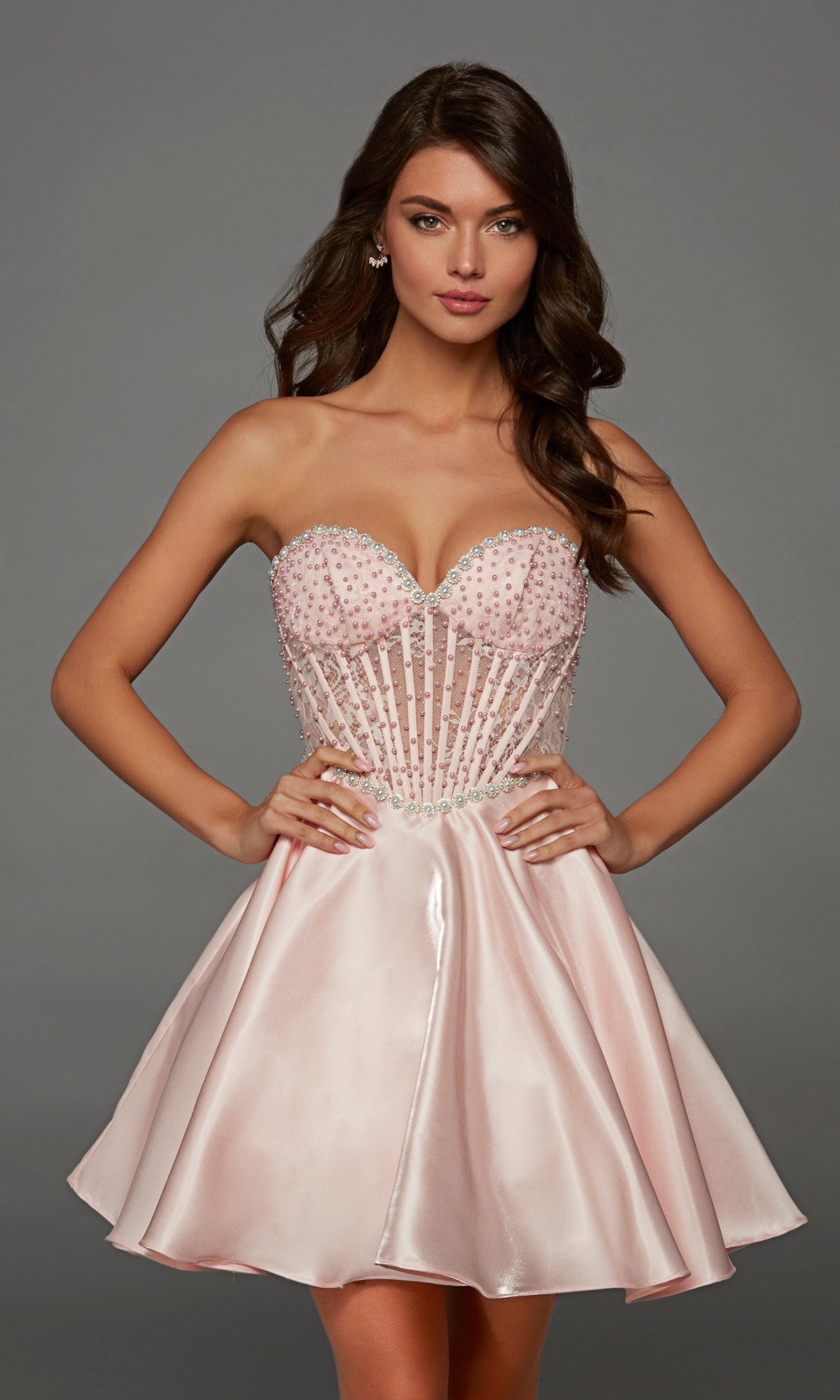 A woman is posing against a plain gray background, wearing the Alyce 30088 short homecoming dress in blush satin, which features a strapless, beaded semi-sheer corset bodice and a flared skirt.