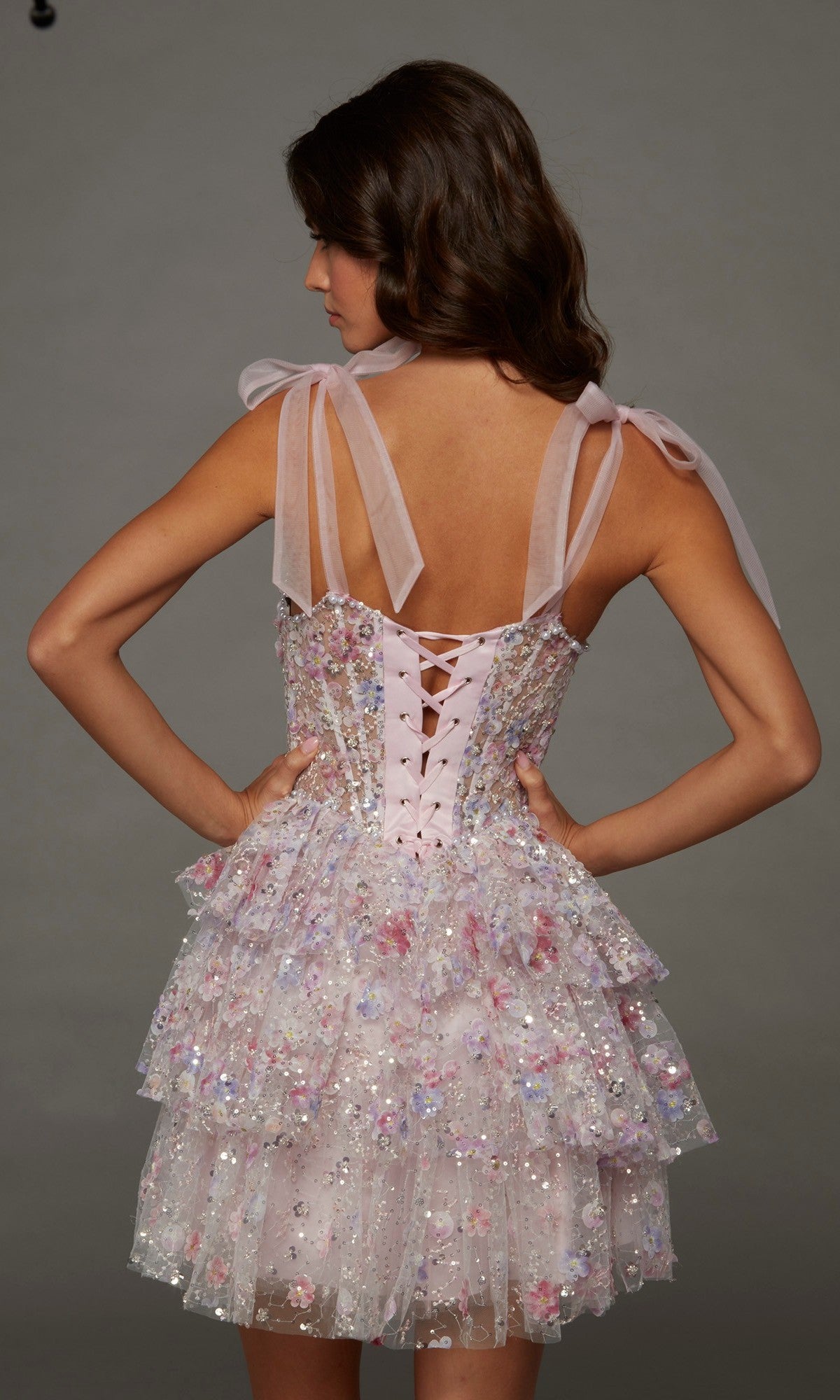 A woman is shown from the back, wearing the Short Homecoming Dress: Alyce 30093—a pastel pink, layered mini dress featuring sequin embellishments, ribbon tie straps, and a corset bodice with a lace-up back—posed against a plain gray background.