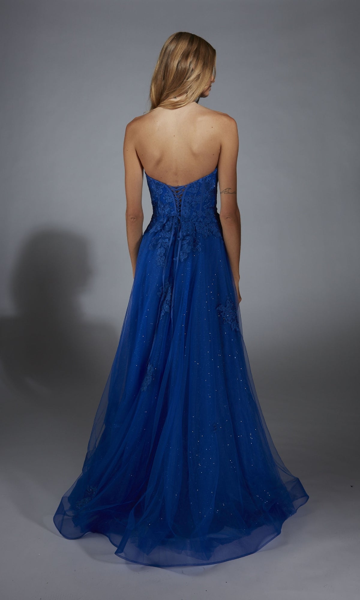 A woman faces away, wearing the Aerie Couture 300 long prom dress—strapless, royal blue, with a fitted corset bodice and a flowing glitter tulle skirt.
