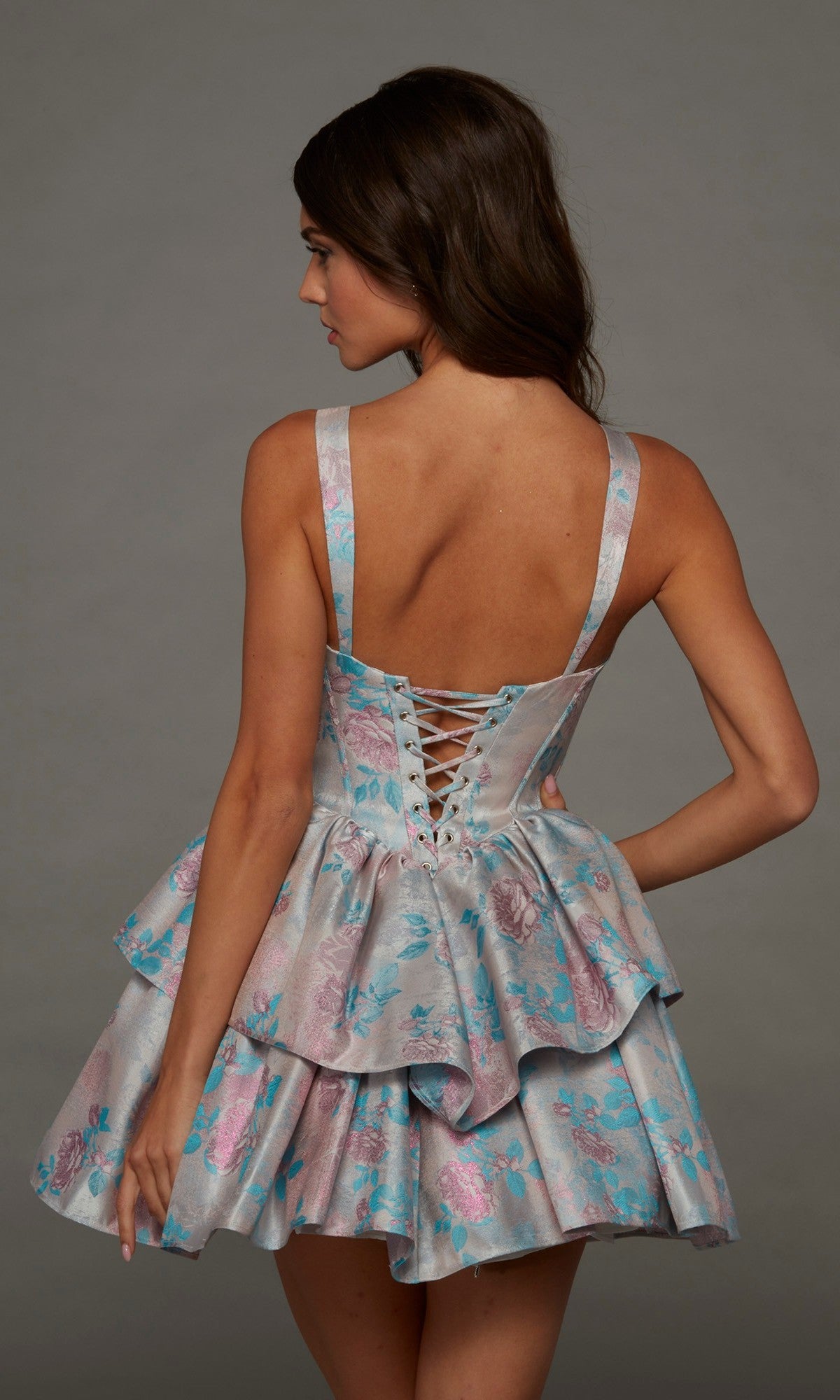 A woman stands against a plain gray background wearing the Alyce 30102 short homecoming dress, which features sleeveless styling, back lacing, ruffled details, and a pastel blue, pink, and silver floral pattern.