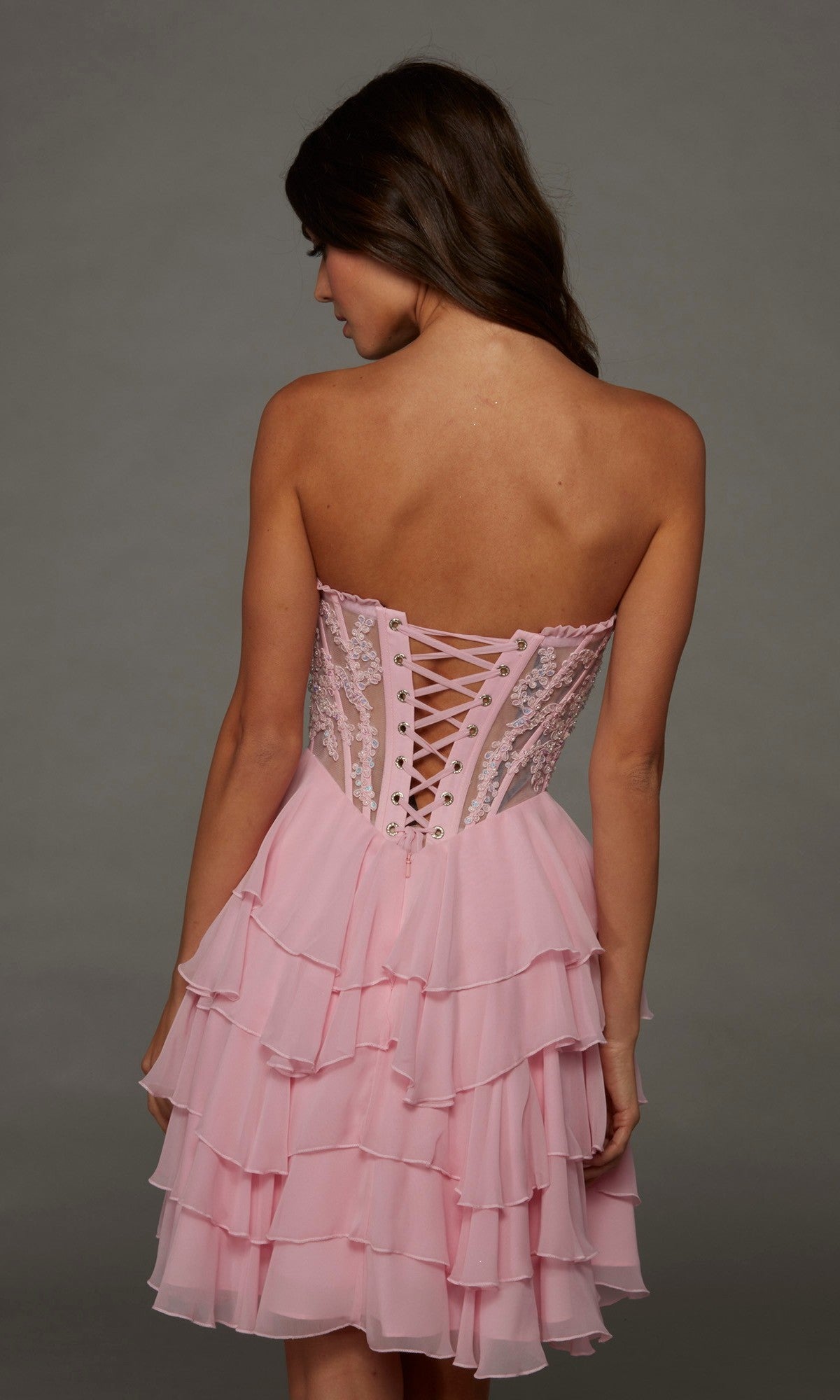 A woman facing away, wearing the Alyce 30104 short homecoming dress featuring a pink strapless corset bodice with a lace-up back and a layered ruffle skirt.
