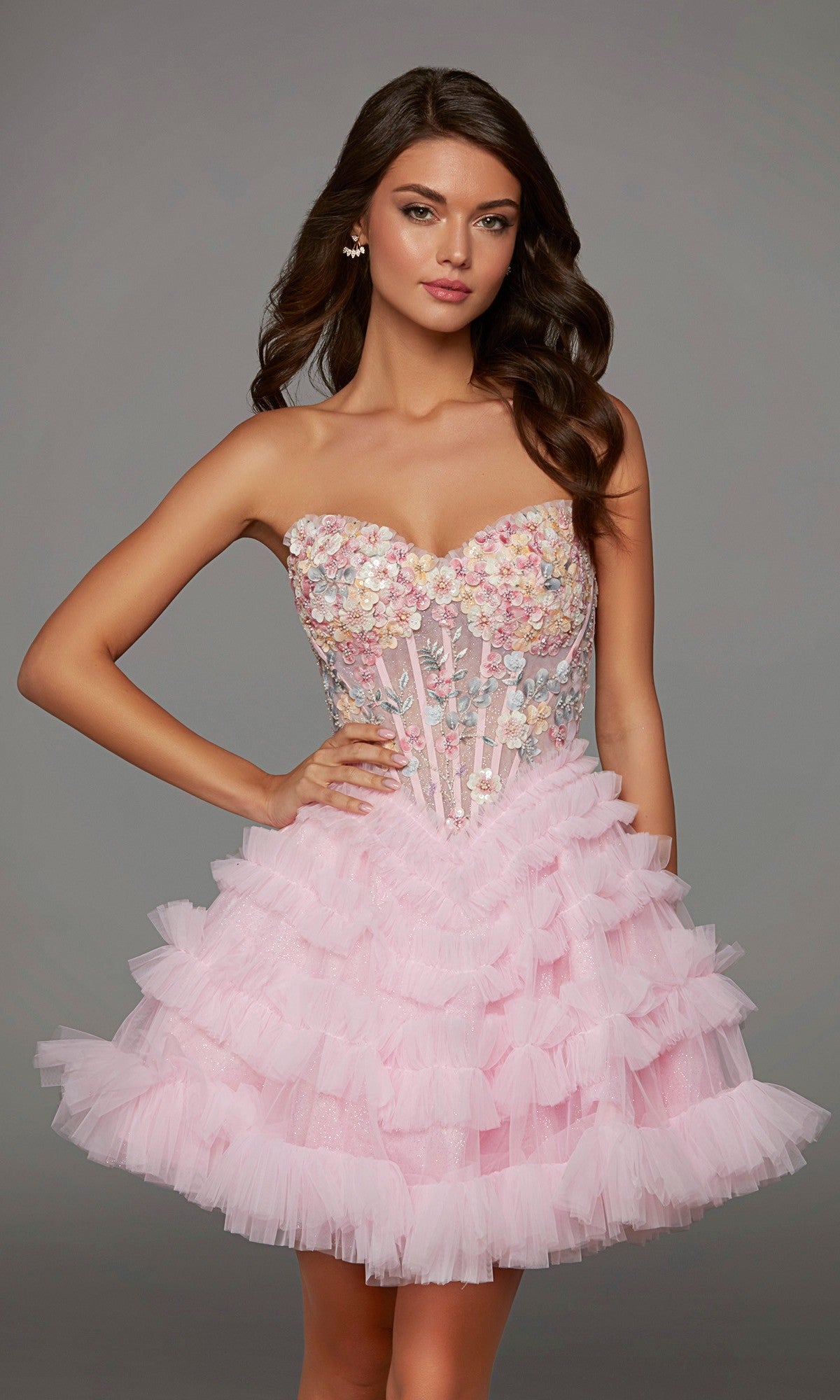A woman stands against a neutral background wearing the Short Homecoming Dress: Alyce 30105 in Rosewater, featuring a pink strapless design with a corset bodice adorned with 3D floral appliques and a layered, ruffled tulle skirt.