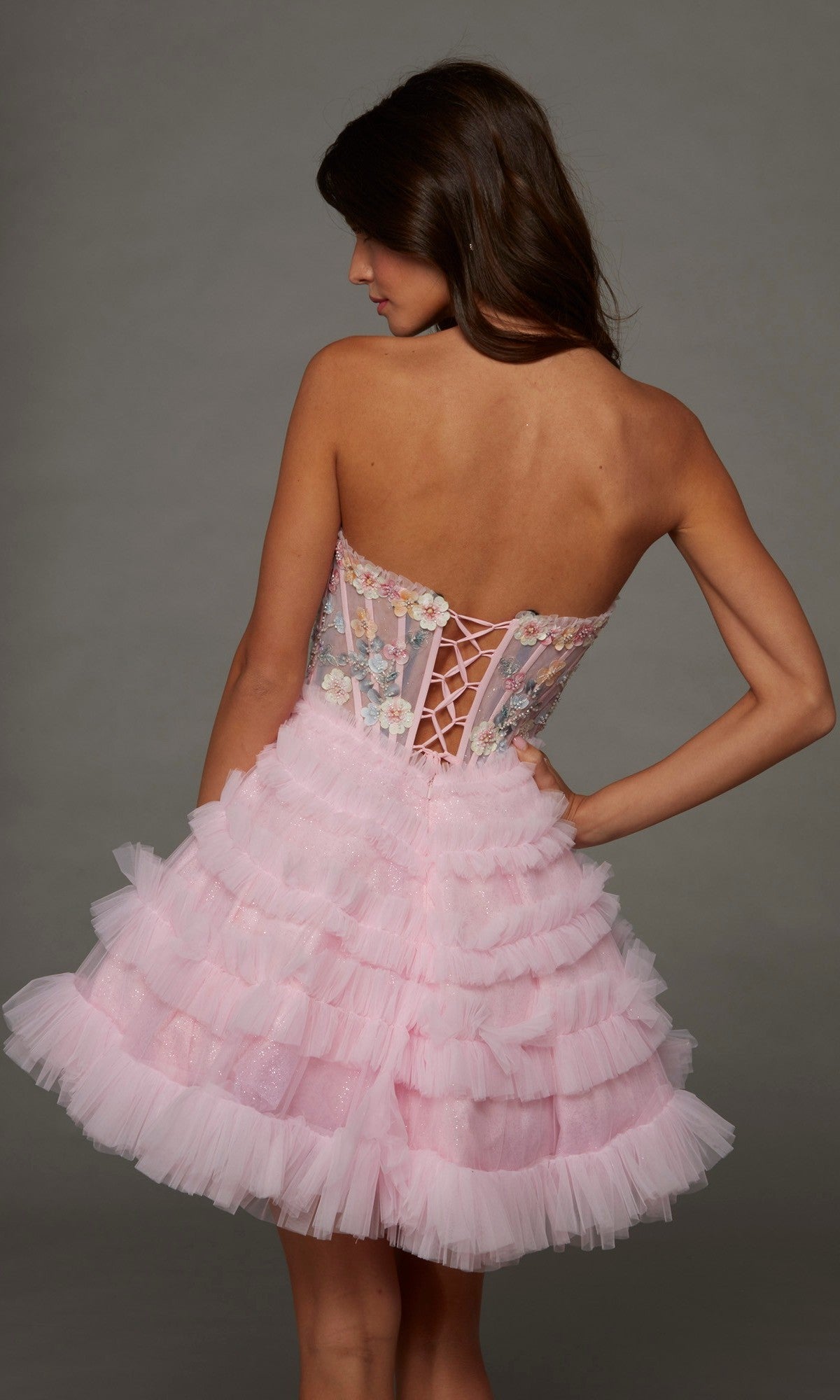 A woman poses with her back to the camera against a gray background, wearing the Alyce 30105 short homecoming dress in pink—a strapless design with a corset bodice adorned with 3D floral appliques and a layered tulle skirt.