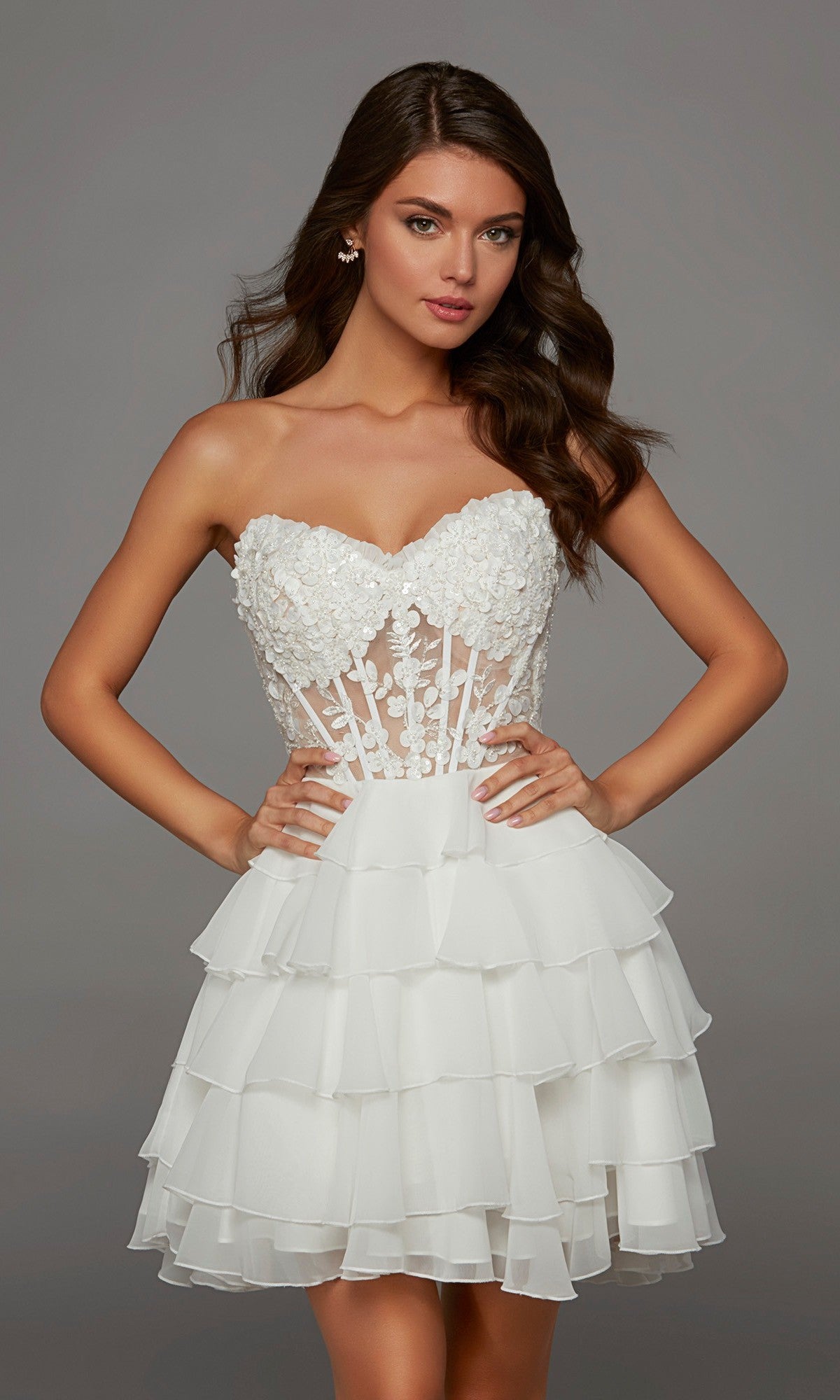 A woman models the Alyce 30109 Short Homecoming Dress in Diamond White, showcasing its strapless design with 3D floral appliques on the bodice and a layered ruffled skirt, as she stands with hands on hips against a plain background.