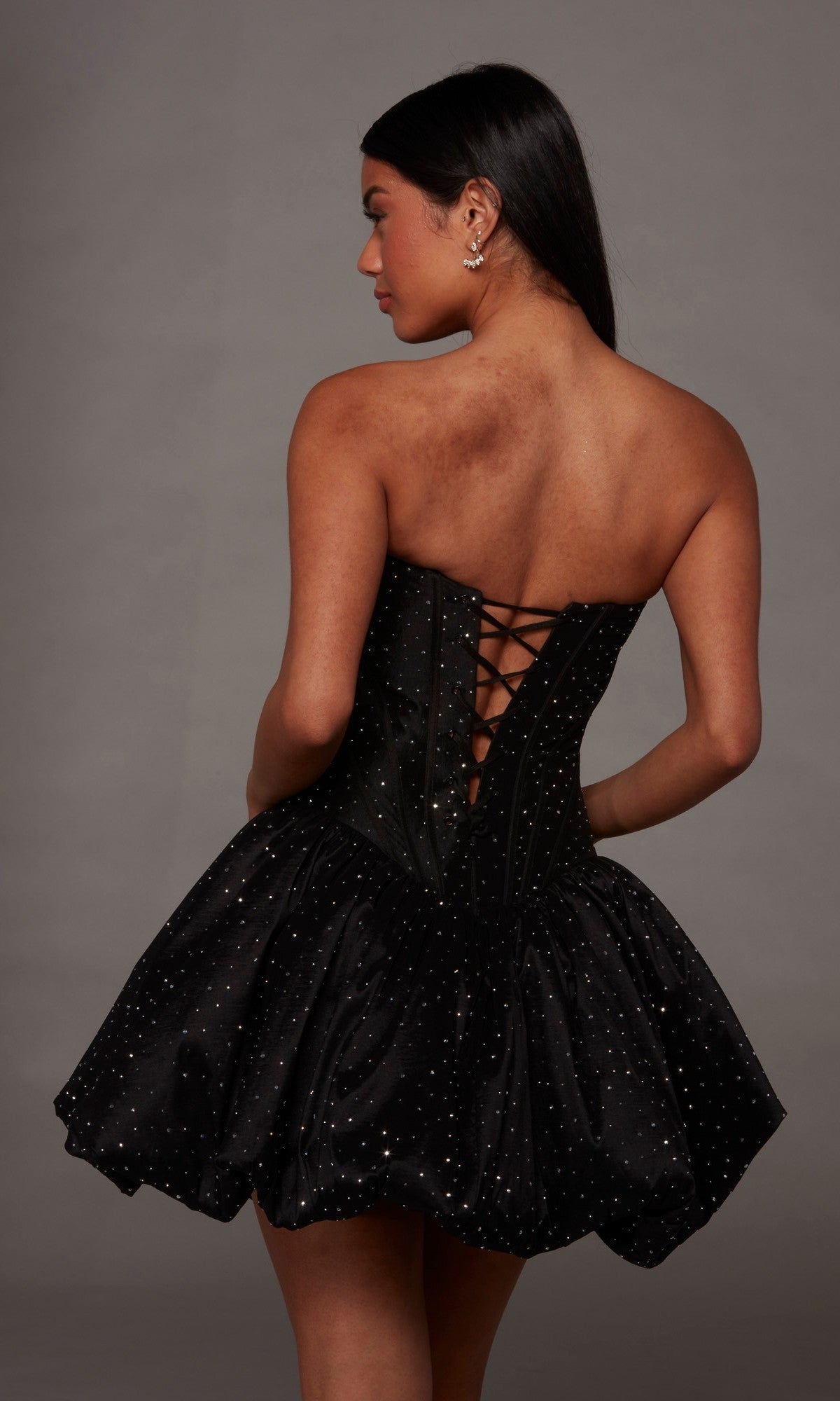 A woman is seen from behind, wearing the Alyce 30125 short homecoming dress, which features a strapless black corset bodice with a lace-up back and sparkling rhinestone accents. The look is completed with a bubble-hem A-line skirt, all set against a simple gray background.