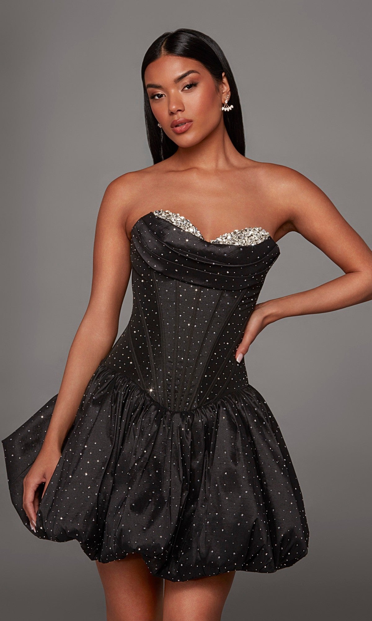 A woman wearing the Alyce 30125 short homecoming dress, featuring a rhinestone strapless fitted bodice and a bubble-hem A-line skirt, stands against a plain gray background.