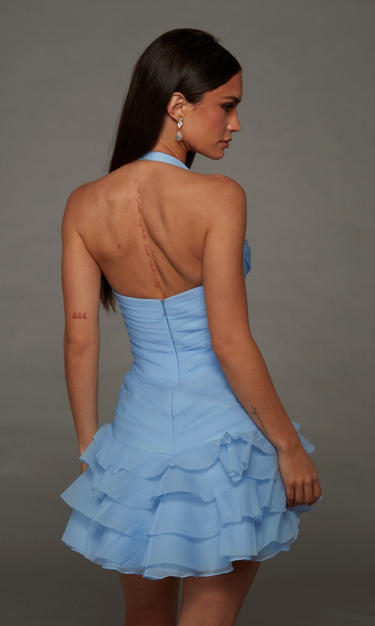 Wearing the Alyce 30127 Short Homecoming Dress in light blue, a woman with long straight hair stands facing away, revealing a vertical back scar and the number 444 on her arm.