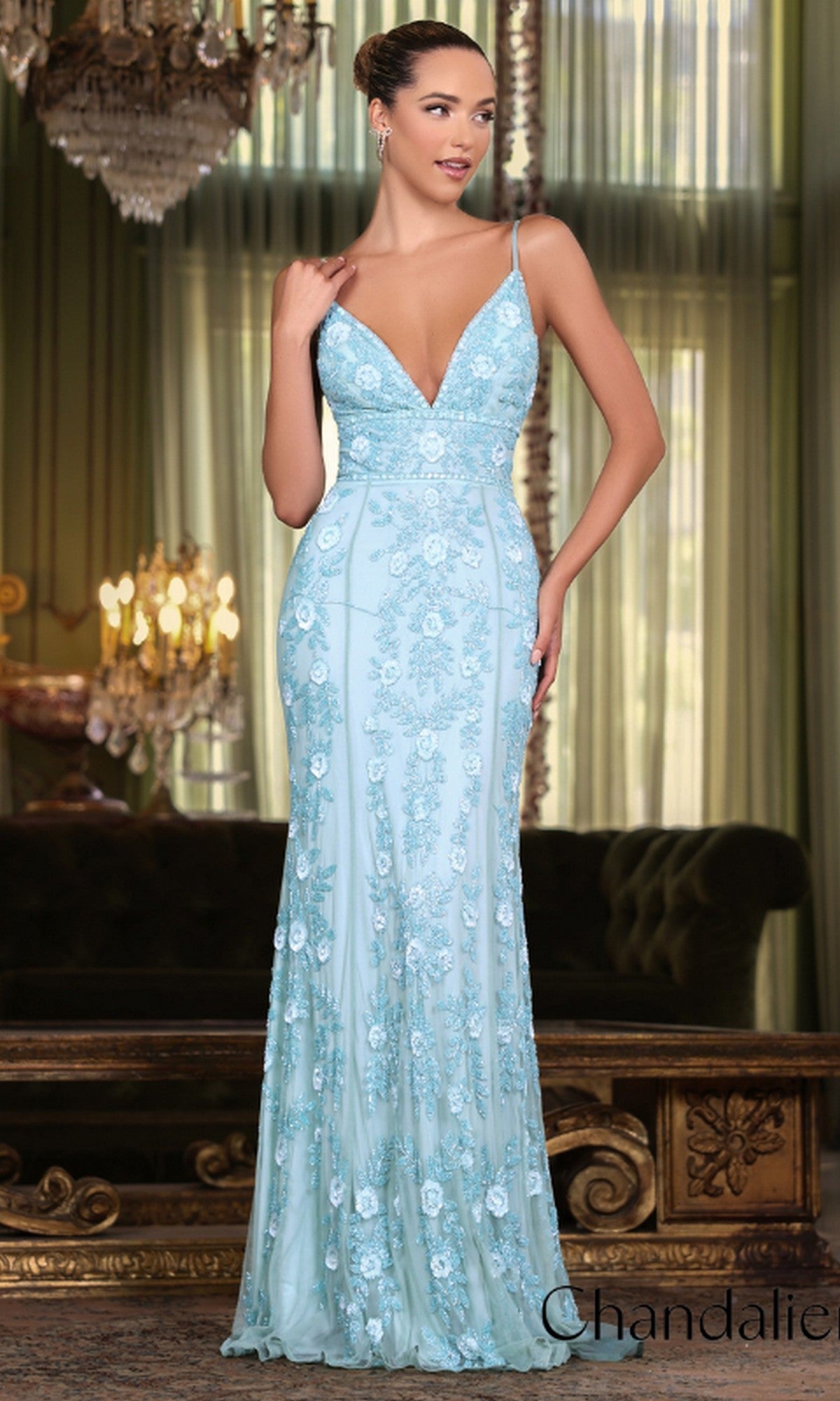 A woman poses indoors in front of a sofa and chandelier, wearing the Long Prom Dress 30157 by Chandalier—a fitted, light blue formal gown with floral beading and thin straps.