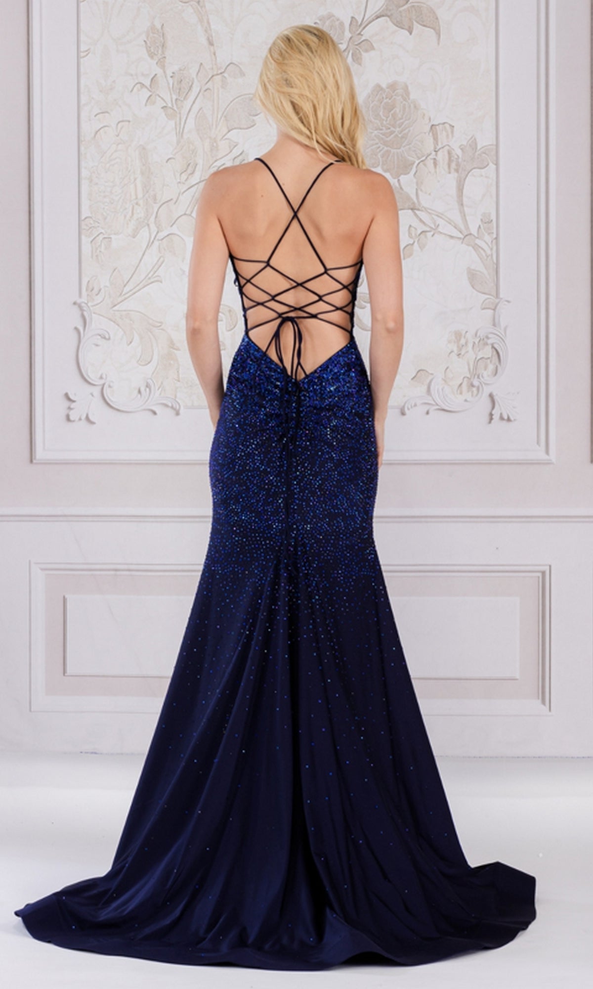 A woman with long blonde hair wears the Open-Back Sparkly Long Beaded Prom Dress 3018, featuring a floor-length silhouette and lace-up open back, standing before a decorative wall.