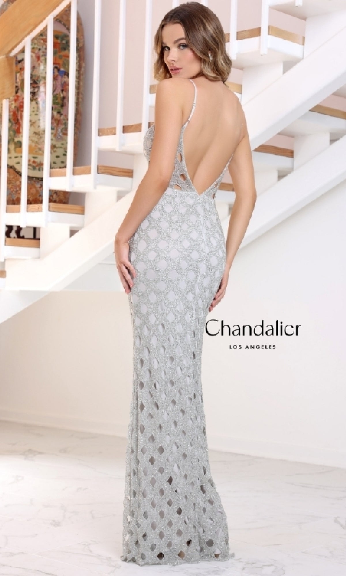 A woman in the Chandalier 30190 long prom dress—silver, sleeveless, with stretch sequins, geometric lace pattern, low back, and a dramatic high-leg slit—stands on a white marble floor before a staircase.