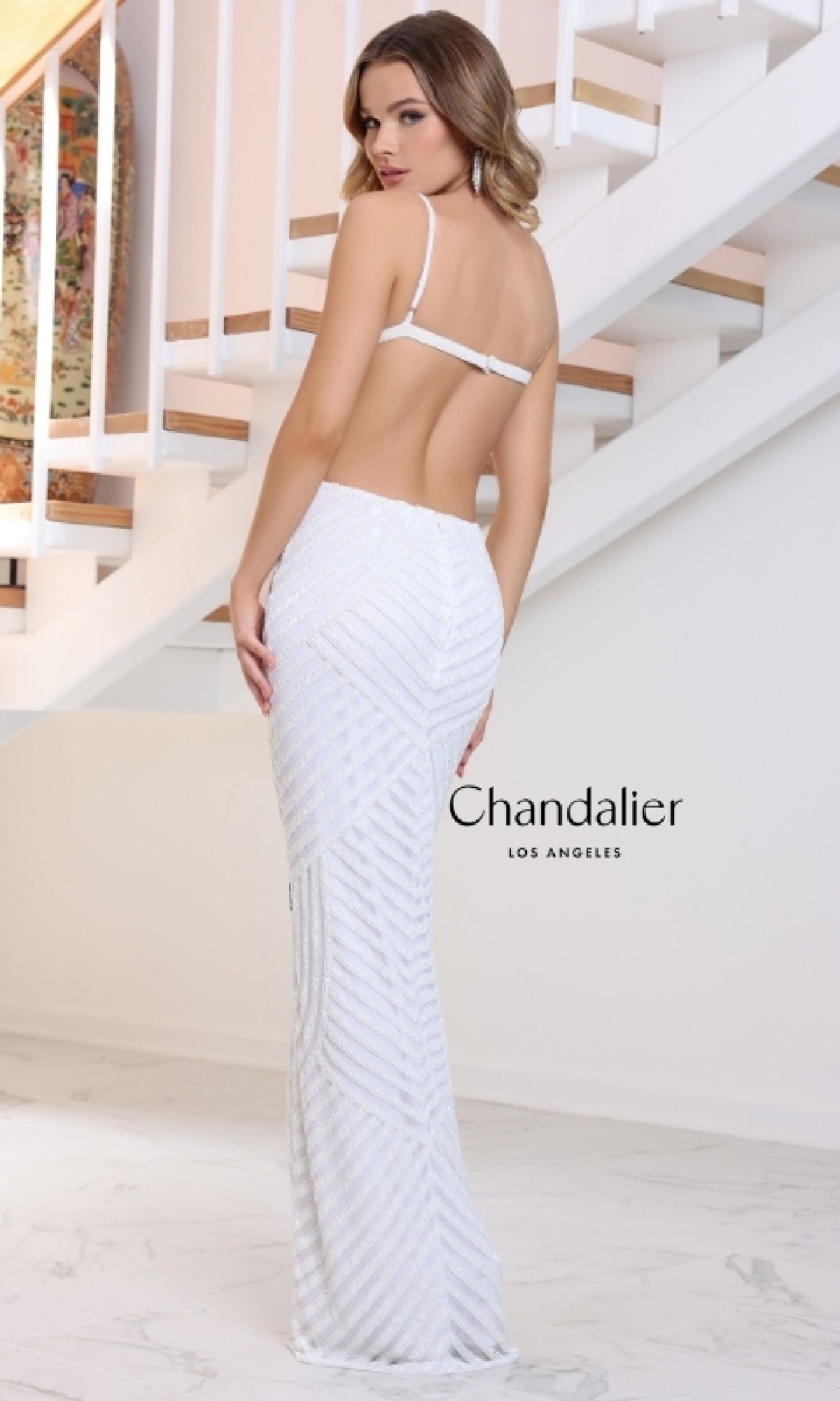 A woman stands indoors near a staircase, facing away in the Long Prom Dress: Chandalier 30211. The white, open back gown features hot stone embellishments.