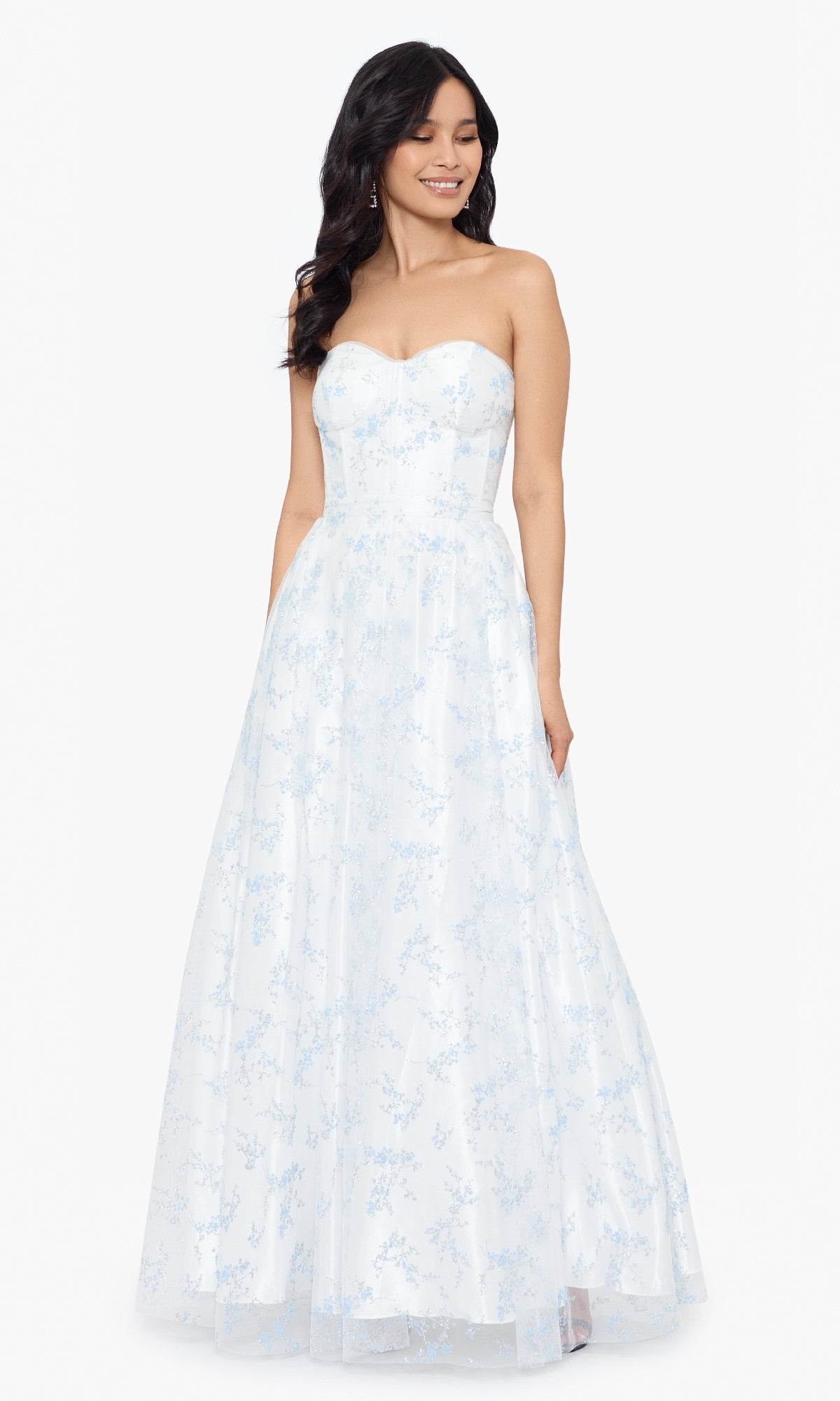 A woman, eyes closed and smiling, stands in the Blondie Nites 3024BN long formal dress—an off-white, strapless gown adorned with a light blue floral pattern.