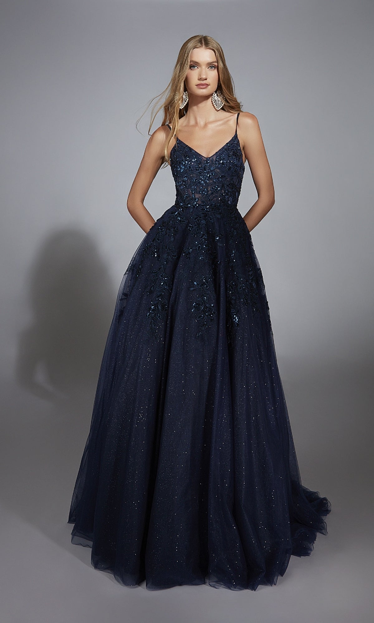 A woman wears the Aerie Couture 302 Long Prom Dress, a navy blue floor-length gown with a fitted v-neck corset bodice, embellished details, and a full tulle skirt, standing against a gray background.