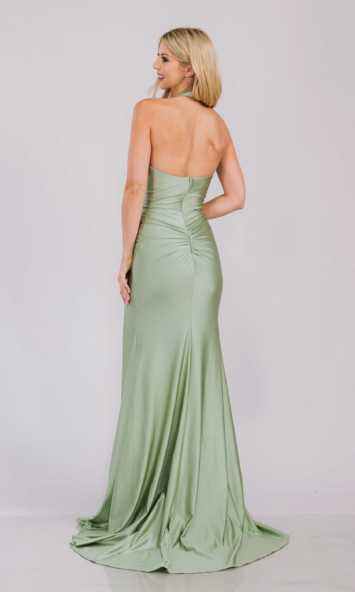 A woman models the V-Neck Long Halter Prom Dress: Amelia Couture 3040, standing with her back to the camera to showcase the ruched detailing and floor-length design against a plain background.