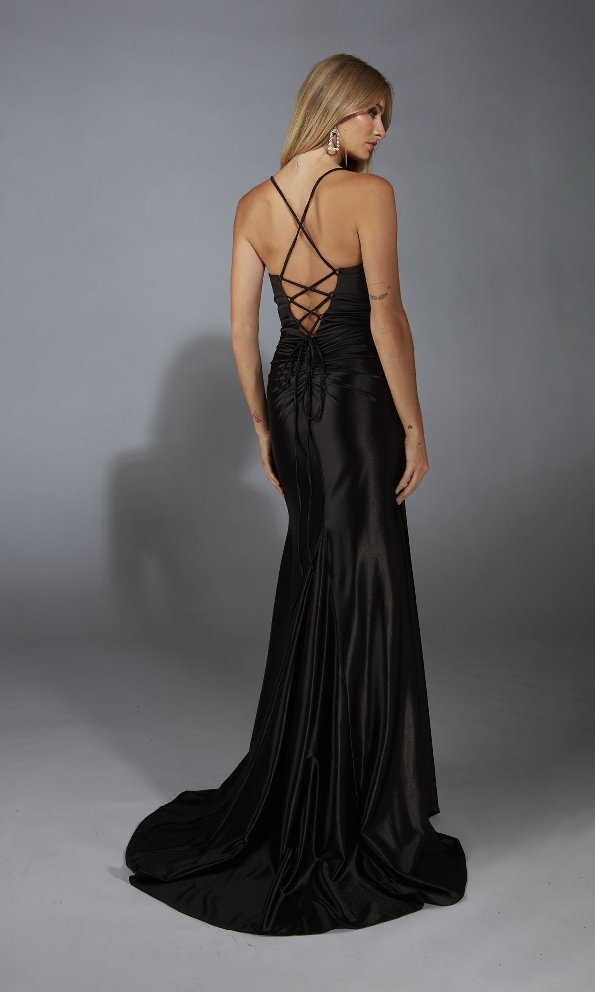 A woman faces away in the Aerie Couture 304, a long black prom dress with a lace-up back, satin fabric, and slight train, set against a plain gray background.