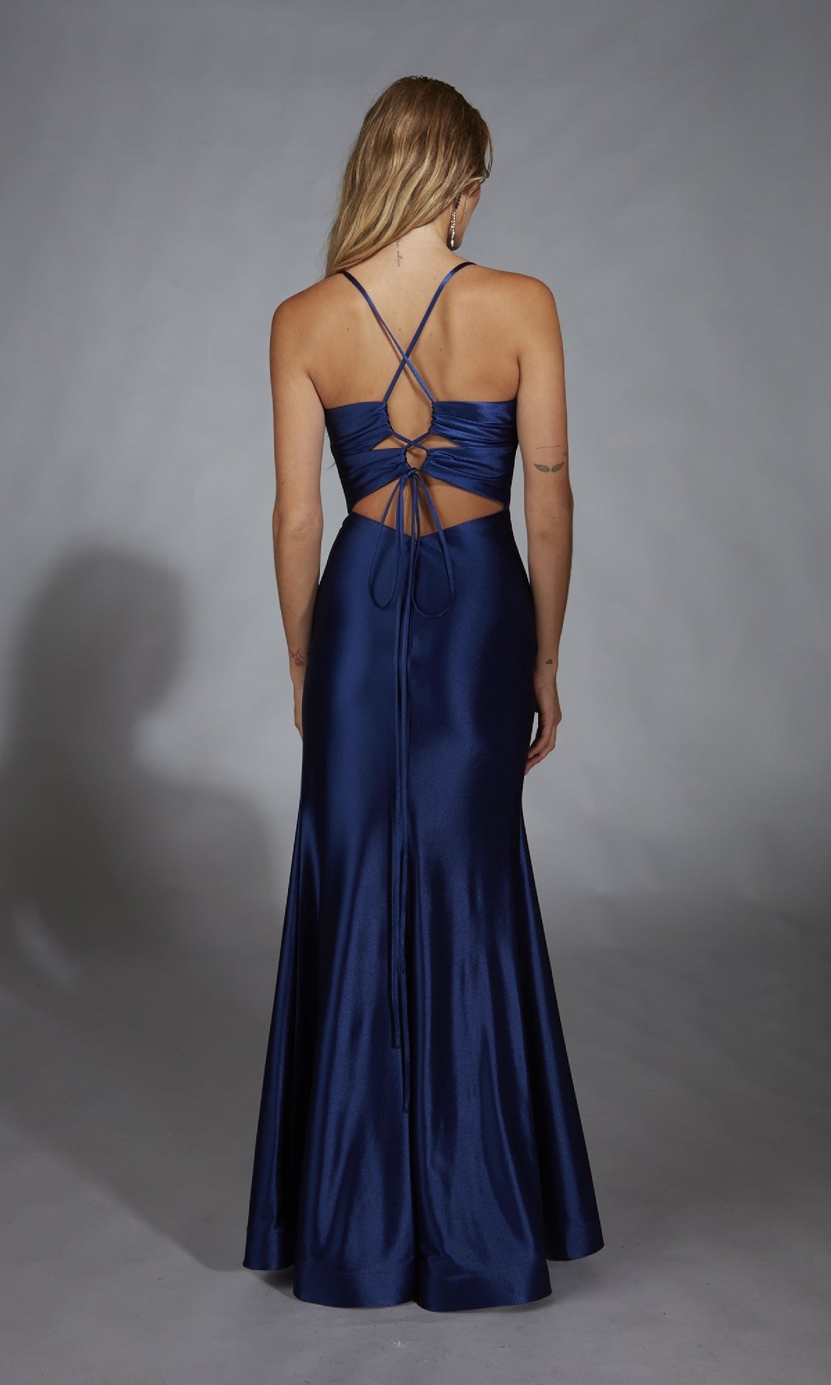 A woman stands facing away against a plain gray background, wearing the Aerie Couture 305 long fitted prom dress in deep blue satin with an open back and lace-up detail.