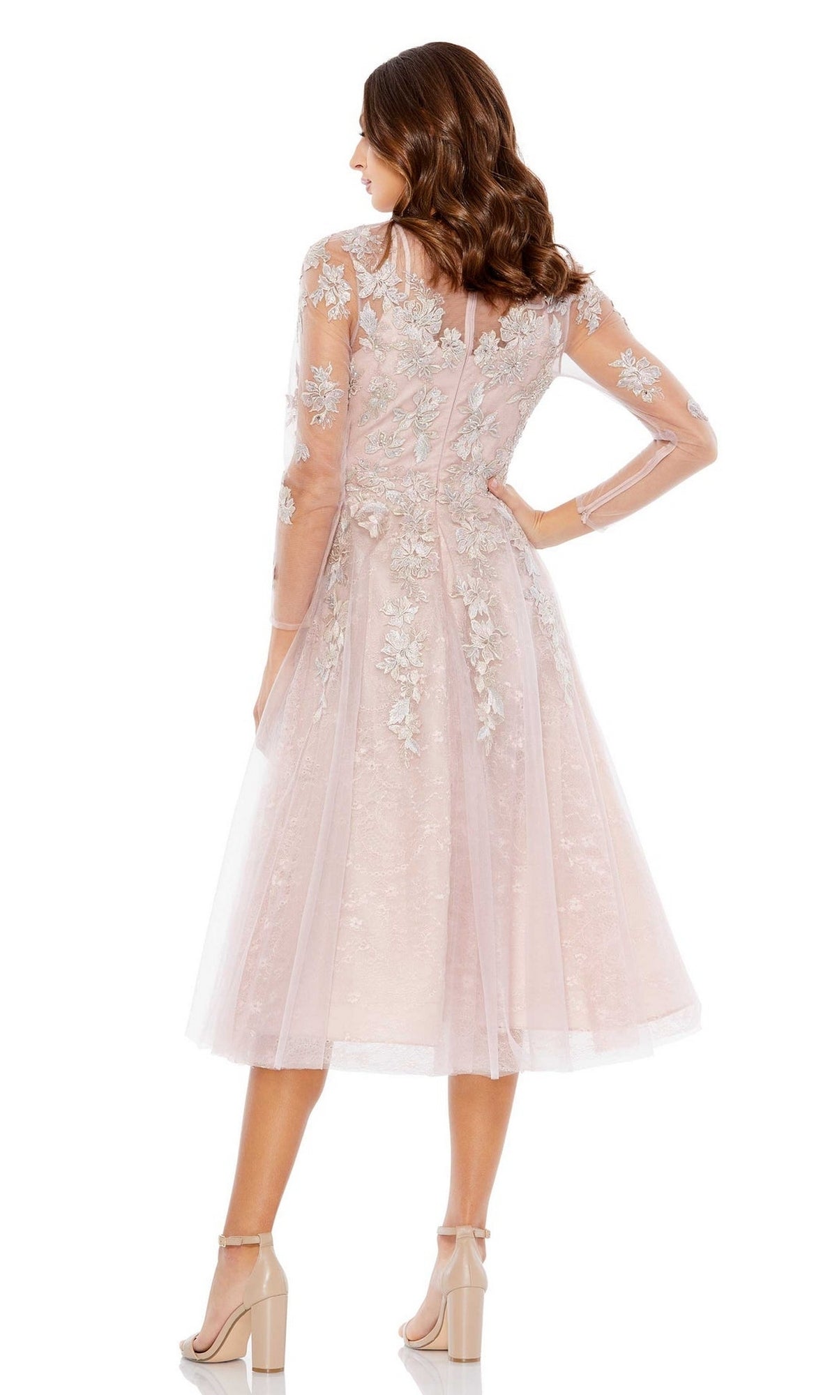 A woman stands with her back to the camera, wearing the Mac Duggal Short Wedding Guest Dress 30681 in light pink with long sheer sleeves, floral embroidery, and nude high-heeled sandals.