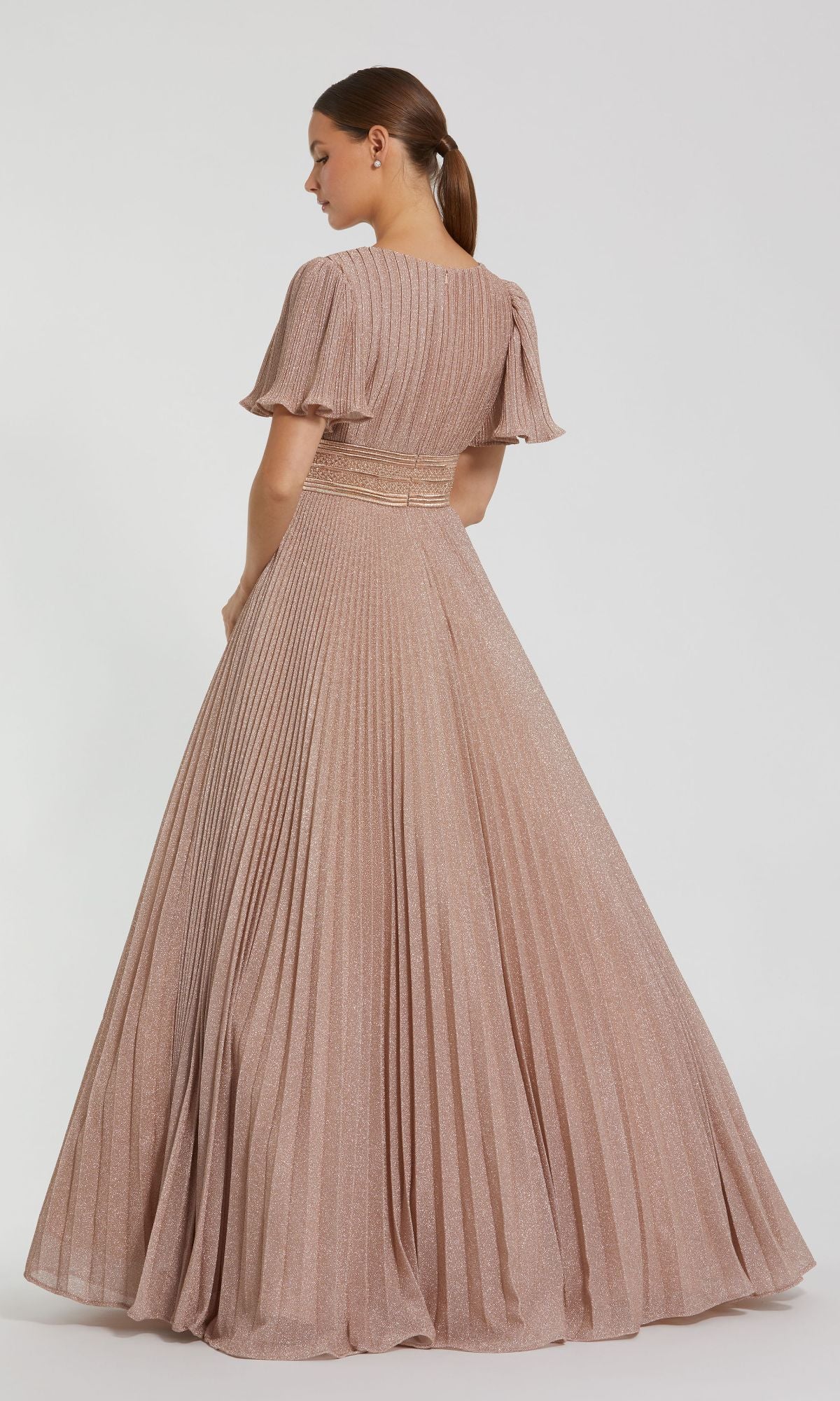 A woman stands facing away against a white background, showcasing the elegance of the Mac Duggal 30776 Short Sleeve Long Formal Dress, a floor-length light pink gown with pleats and flutter sleeves.
