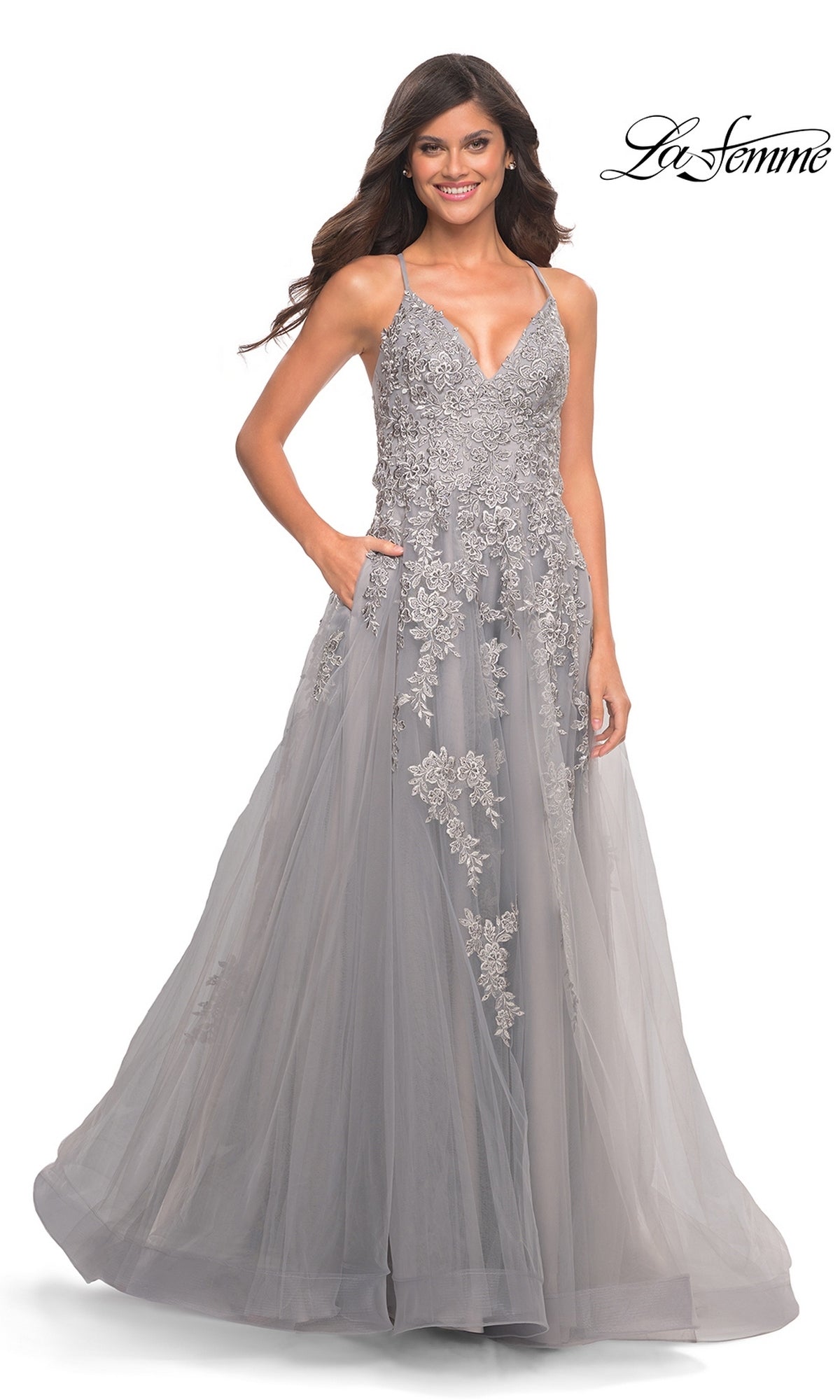 A woman wears a sleeveless, floor-length gray La Femme 30810 prom dress with a V-neckline, embroidered floral appliqués on the bodice, and a full tulle skirt with pockets.