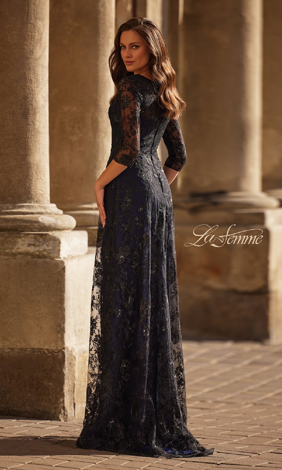 A woman in the Black and Blue Lace Formal Gown: La Femme 30825, featuring sheer sleeves and floor-length lace, stands outdoors near stone columns, facing slightly away from the camera. The detailed gown is both elegant and timeless.