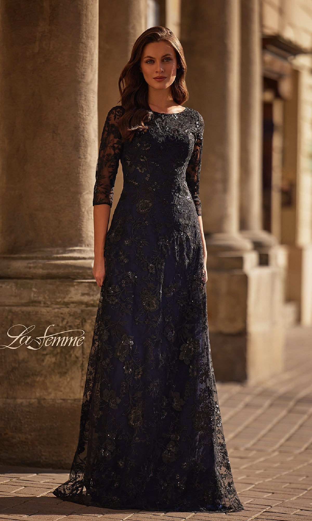 A woman in the Black and Blue Lace Formal Gown: La Femme 30825, featuring three-quarter sleeves, stands outdoors before stone columns.