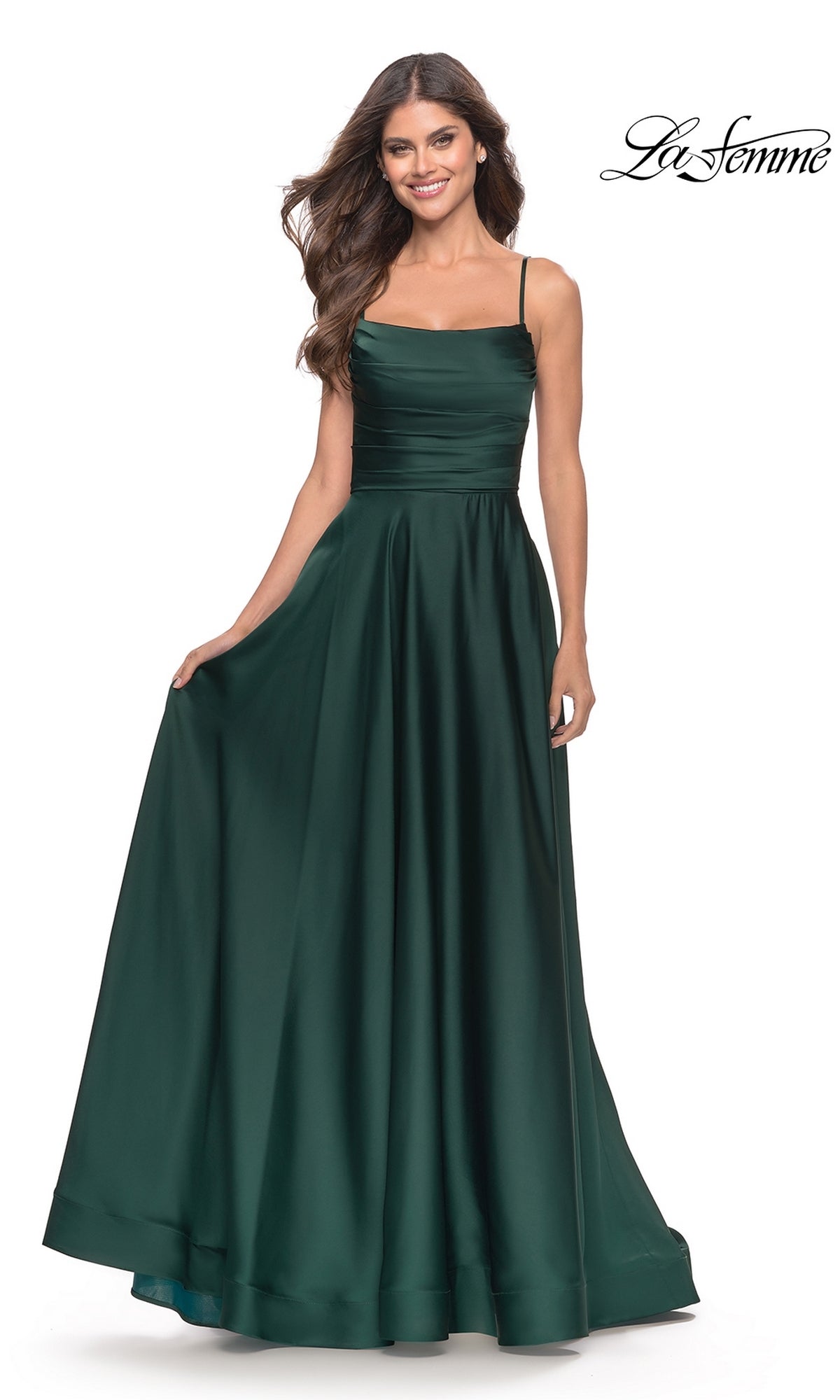 Woman in a sleeveless dark green satin Corset-Back La Femme Long A-Line Prom Dress 31105 with fitted bodice, full skirt, and thin straps, standing while holding out the side of the gown.