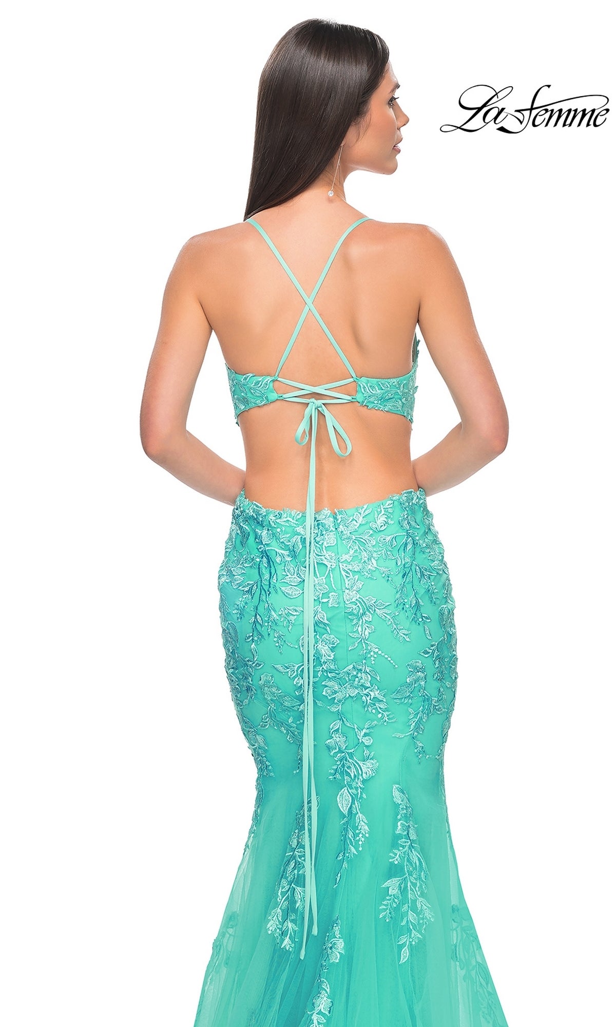 A woman models the La Femme Long Lace Mermaid Prom Dress 31133, featuring turquoise floral embroidery, a backless mermaid silhouette, and crisscross straps against a white background.