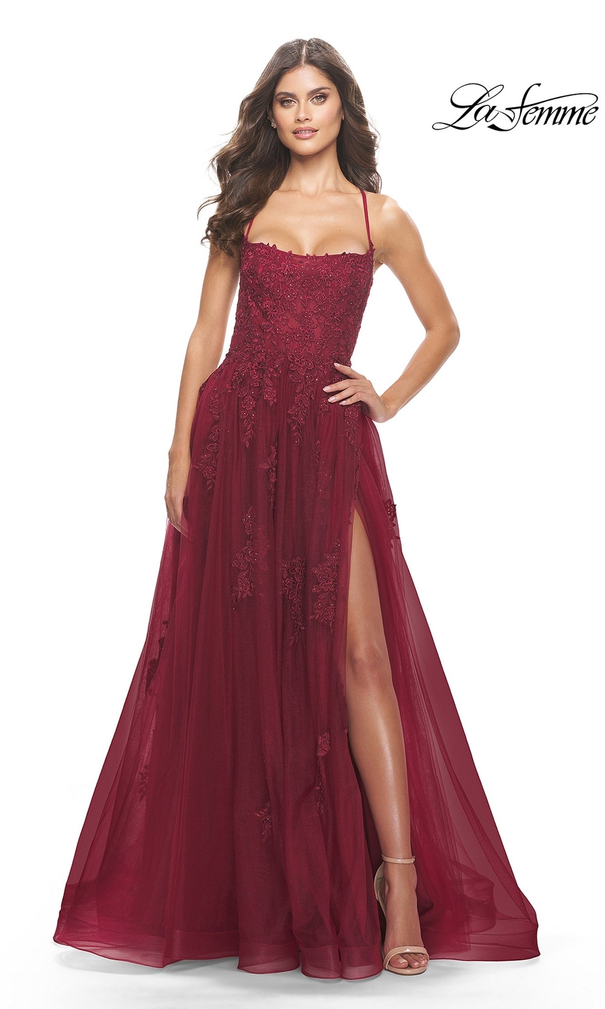 A woman stands against a white background in the La Femme Long A-Line Lace Prom Ball Gown 31135, featuring thin straps, a structured bodice, sleeveless design, floor-length embroidered lace with a high slit, and nude heels.