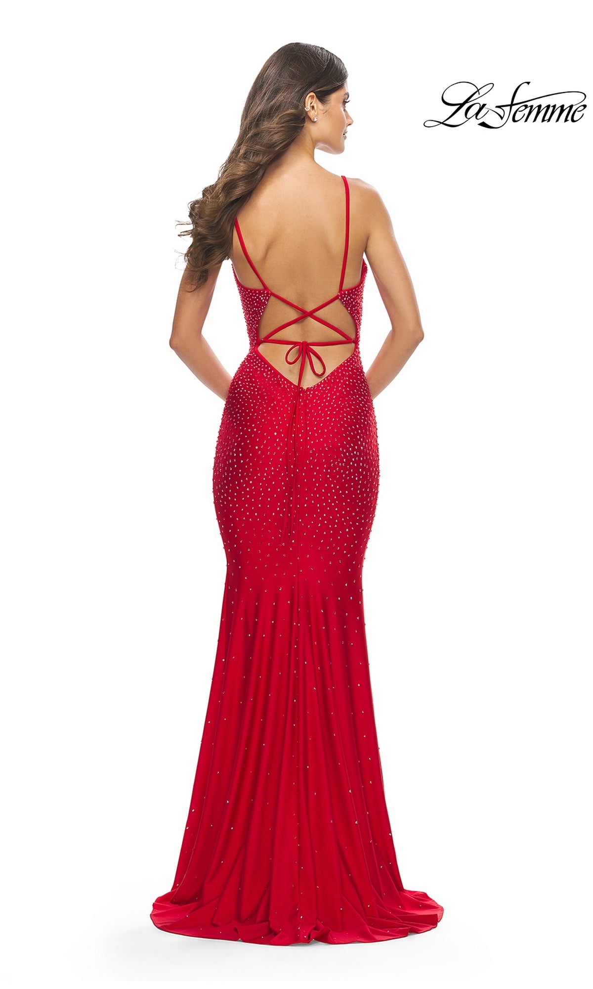 A woman in the La Femme Backless V-Neck Jersey Prom Dress 31215—red, floor-length, with thin straps, a crisscross lace-up back, and subtle embellishments—stands against a white background.