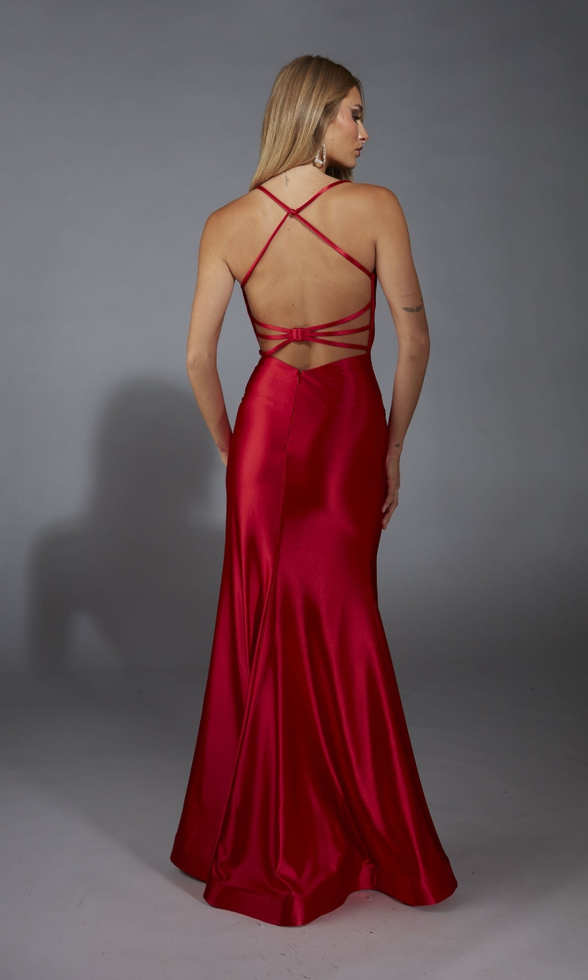 A woman wearing the Aerie Couture 312 long prom dress, featuring red satin fabric and a strappy open back, stands facing away against a plain gray background.