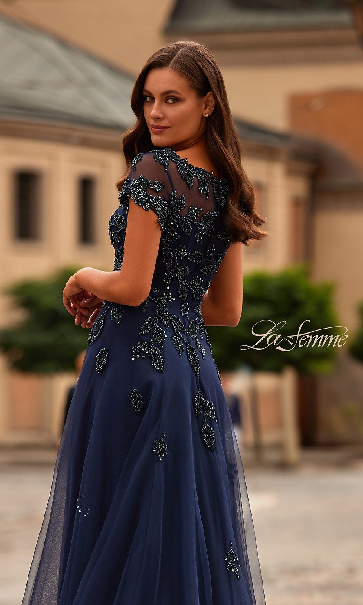 A woman outdoors wears the La Femme 31692, a short sleeve navy formal dress with floral lace and bead detailing on sheer fabric, glancing over her shoulder.