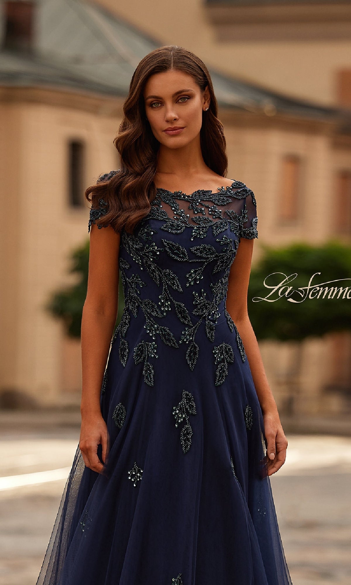 A woman stands outdoors in front of a building, wearing the La Femme 31692 short sleeve navy formal dress with leaf and bead embellishments.
