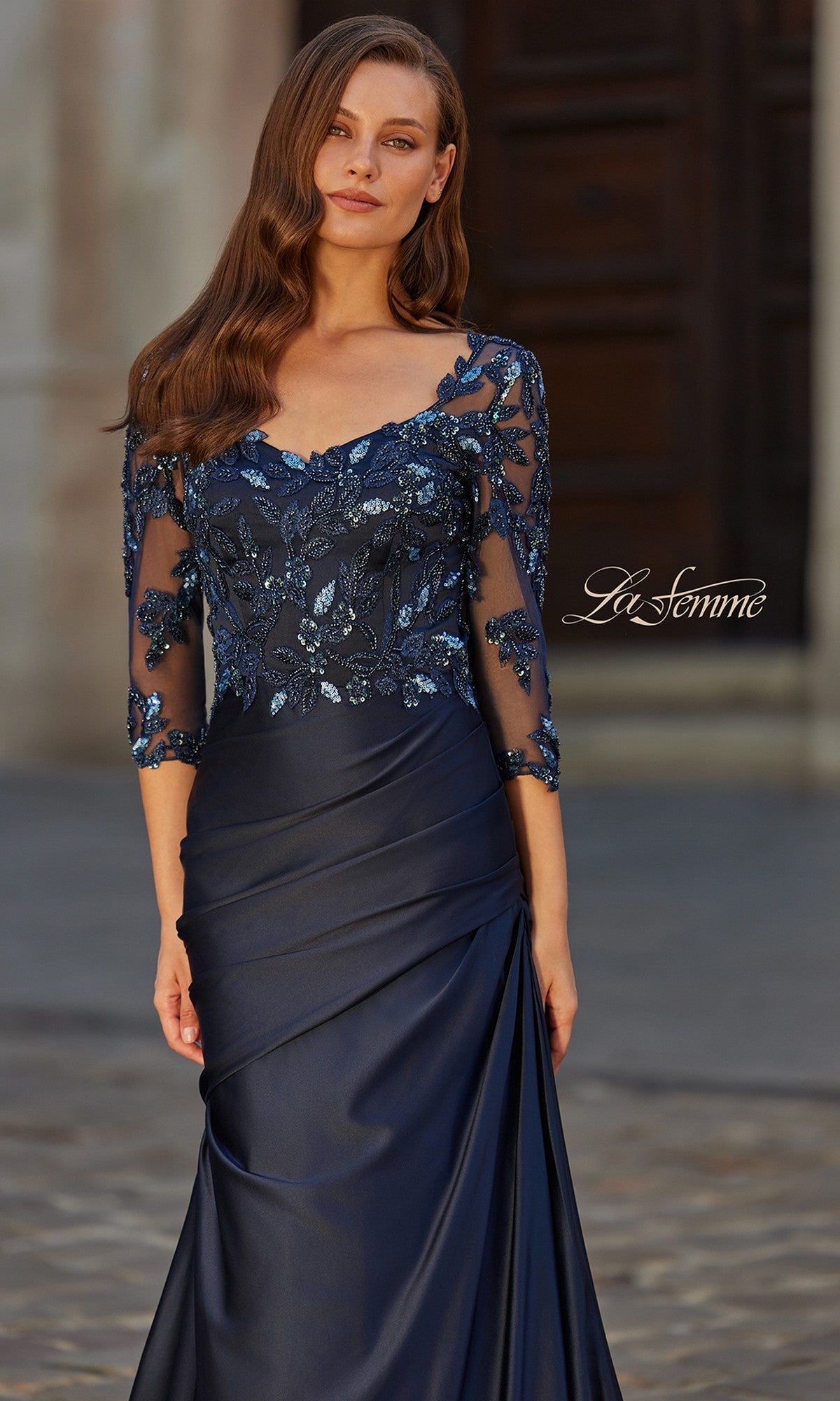 A woman stands outdoors on a cobblestone path wearing the La Femme 31802 long formal dress—a navy blue satin gown with a sheer, sequined beaded lace bodice and three-quarter sleeves.