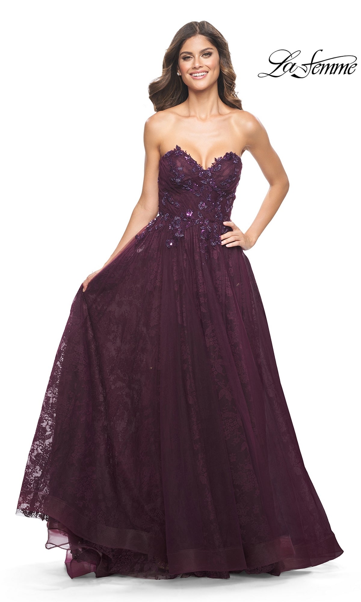 A woman smiles against a white background, wearing the La Femme Strapless Long Prom Ball Gown 31954—a floor-length deep purple gown featuring a sweetheart neckline, lace details, and floral embellishments.