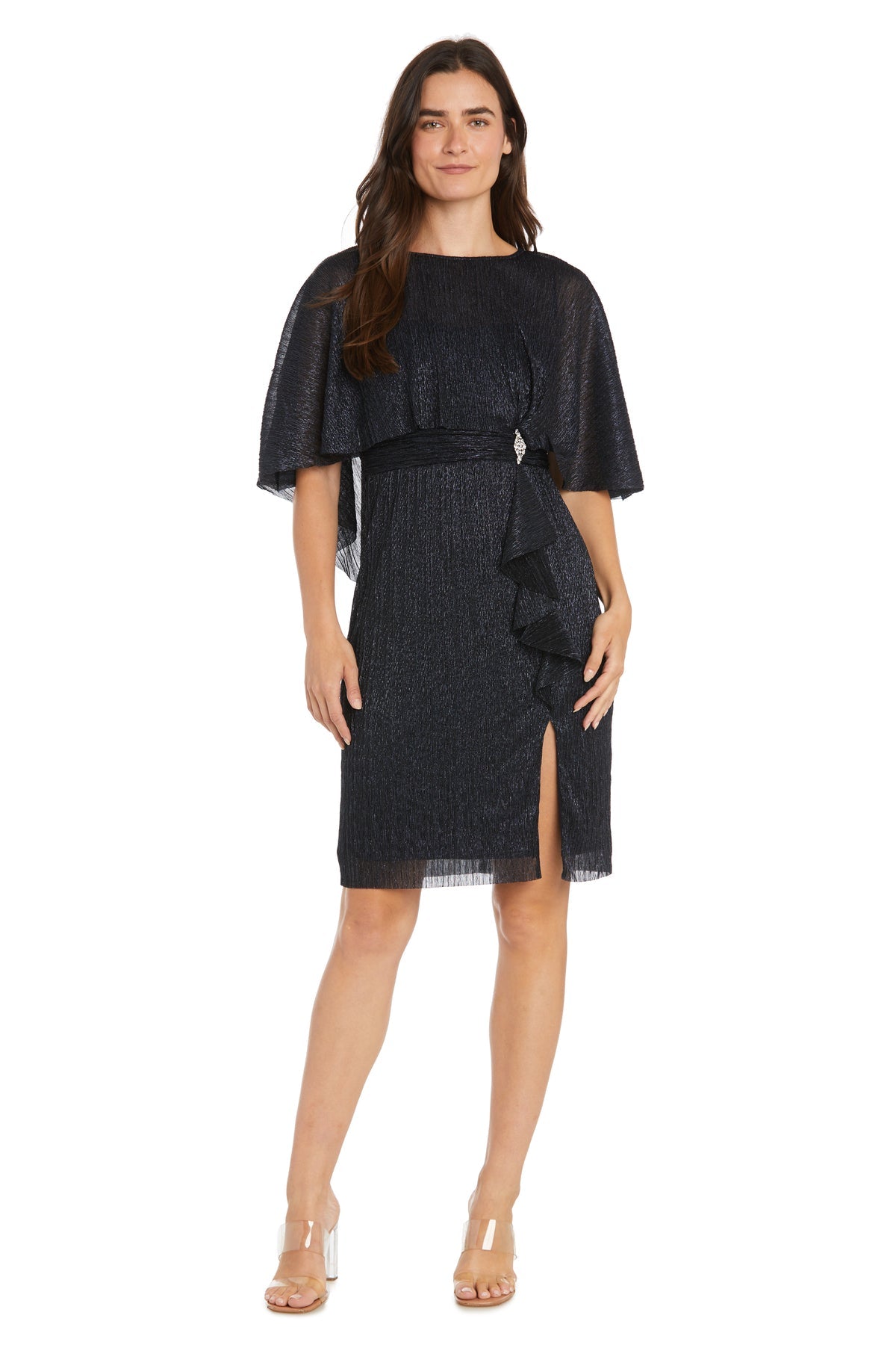 A woman wears the R&M Richards 3195P Caped Knee-Length Party Dress with flutter sleeves, ruffle detail, and a front slit, paired with clear-heeled sandals.