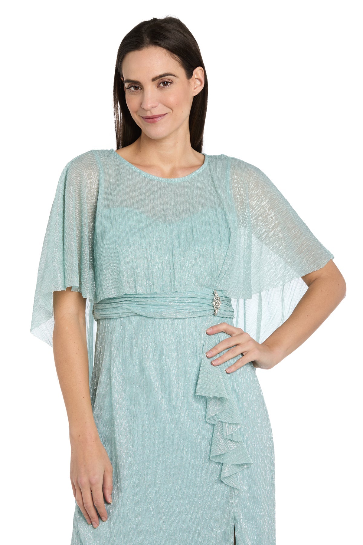 A woman in the R&M Richards 3195P Caped Knee-Length Party Dress, featuring light blue textured fabric, loose short sleeves, and a decorative waist detail, stands with one hand on her hip.