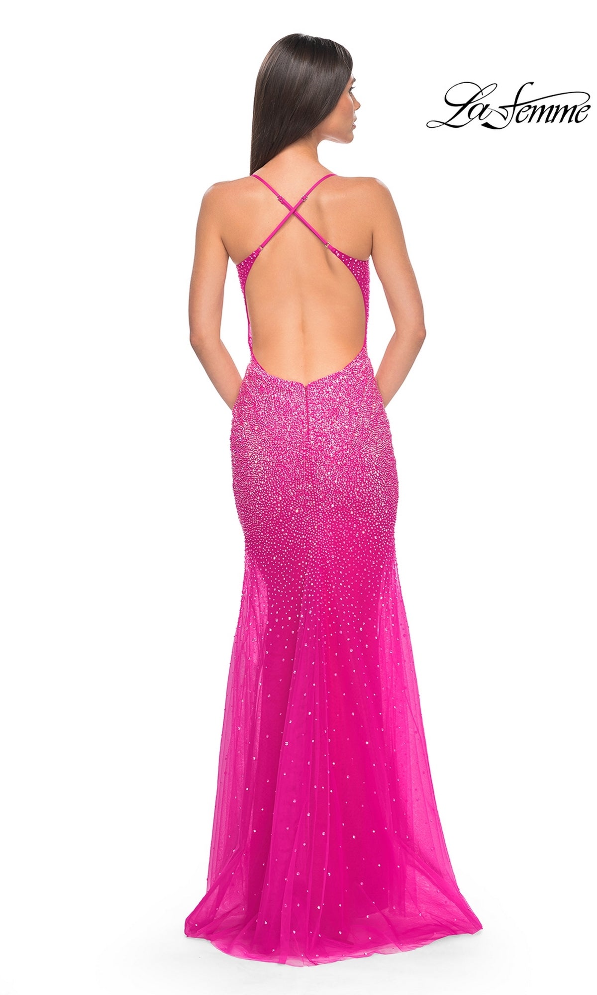 A woman facing away wears the La Femme Sheer-Waist Long Beaded Prom Dress 32007, a bright pink, floor-length, backless gown featuring crisscross straps and sparkling rhinestone embellishments.