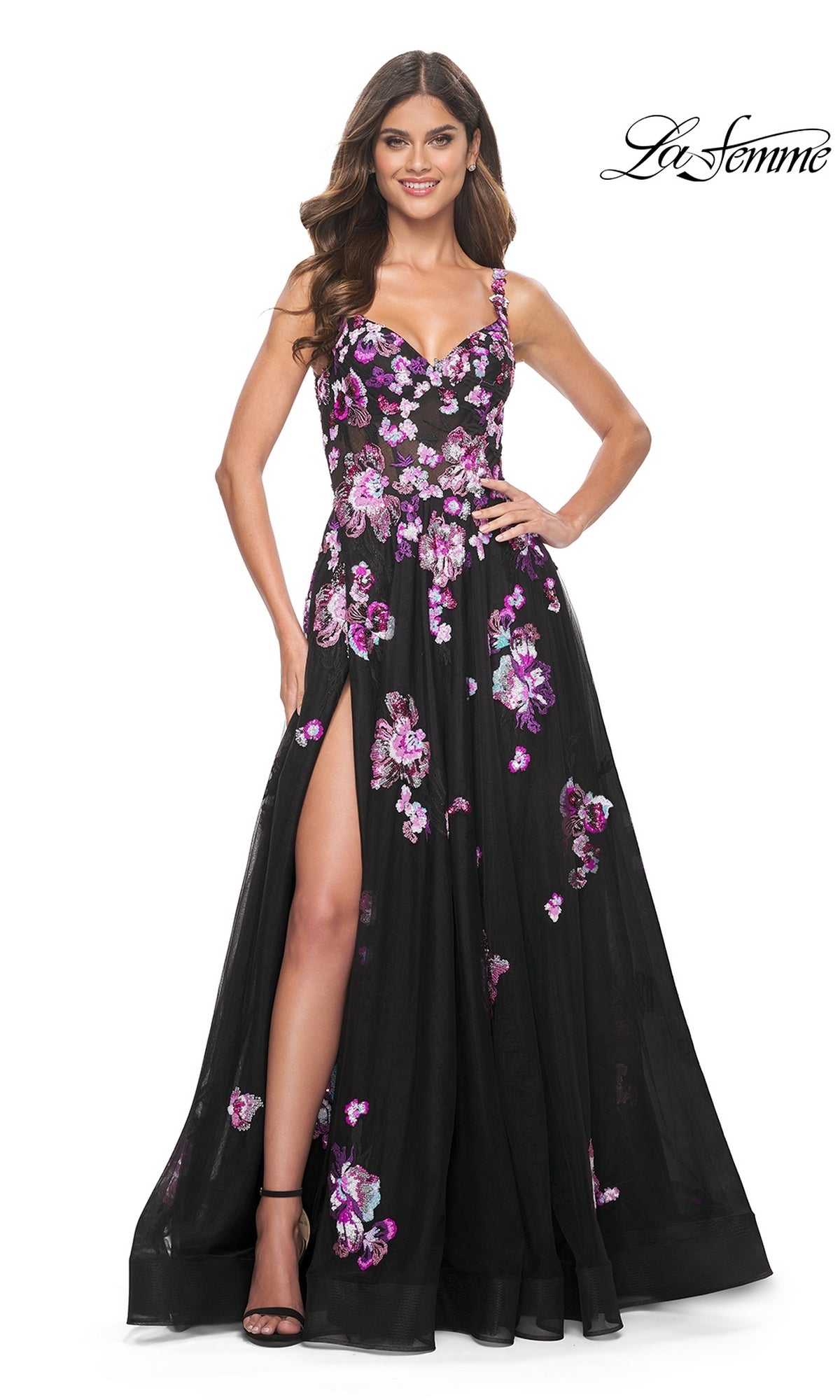 A woman in the La Femme Purple-Floral Long Black Prom Dress 32030, featuring purple sequin flowers, a thigh-high slit, and spaghetti straps, stands smiling with one hand on her hip.