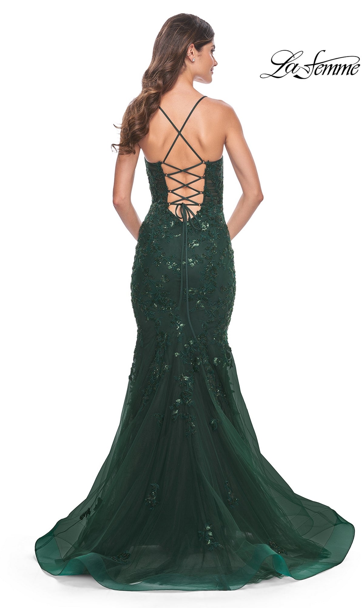A woman stands with her back to the camera, wearing the La Femme Tonal Lace Long Mermaid Prom Dress 32033 in dark green, featuring beaded lace embellishments and a lace-up open back.