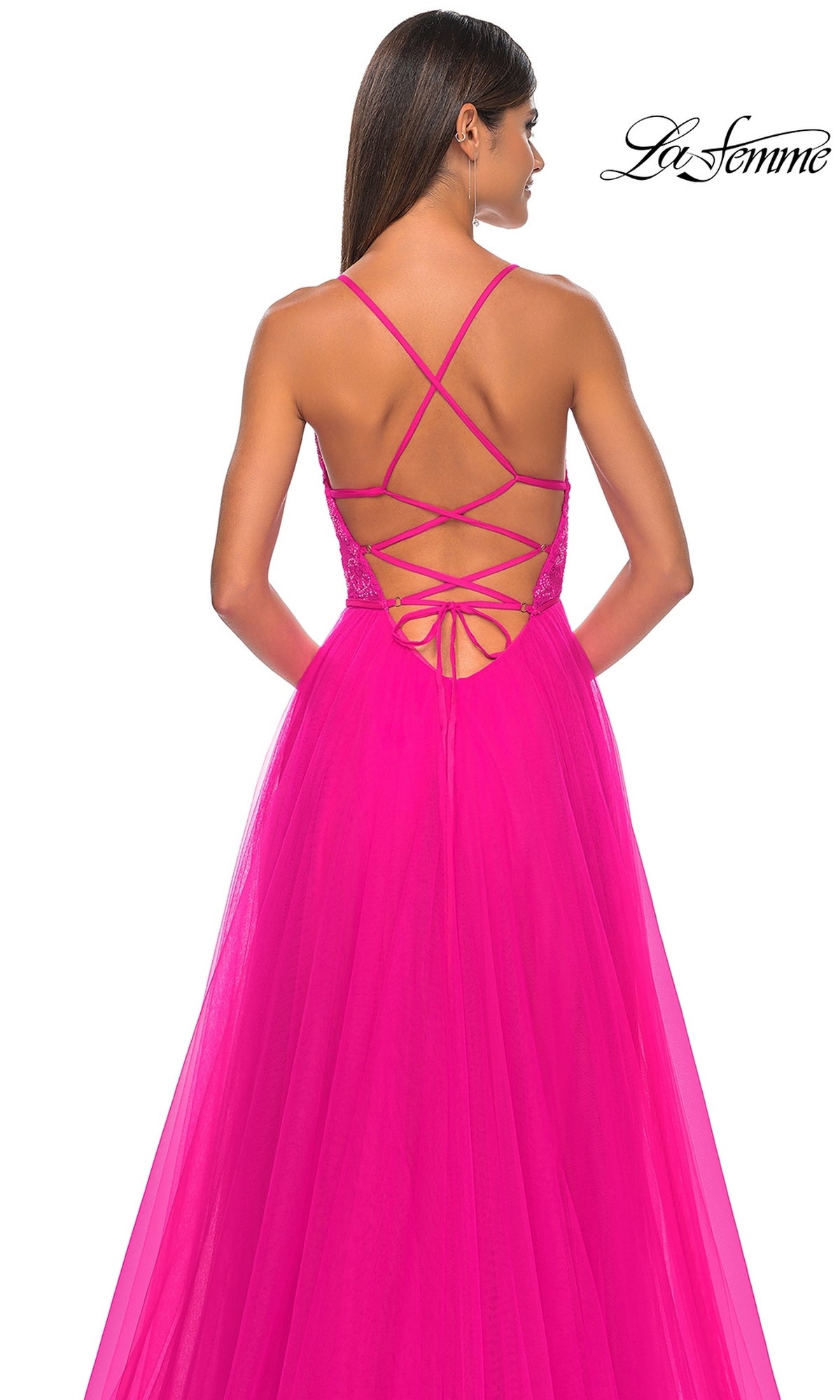 A woman stands facing away, wearing the La Femme Deep V-Neck Long Tulle Prom Gown 32059—a bright pink, floor-length A-line dress with a crisscross strappy open back and long tulle skirt.