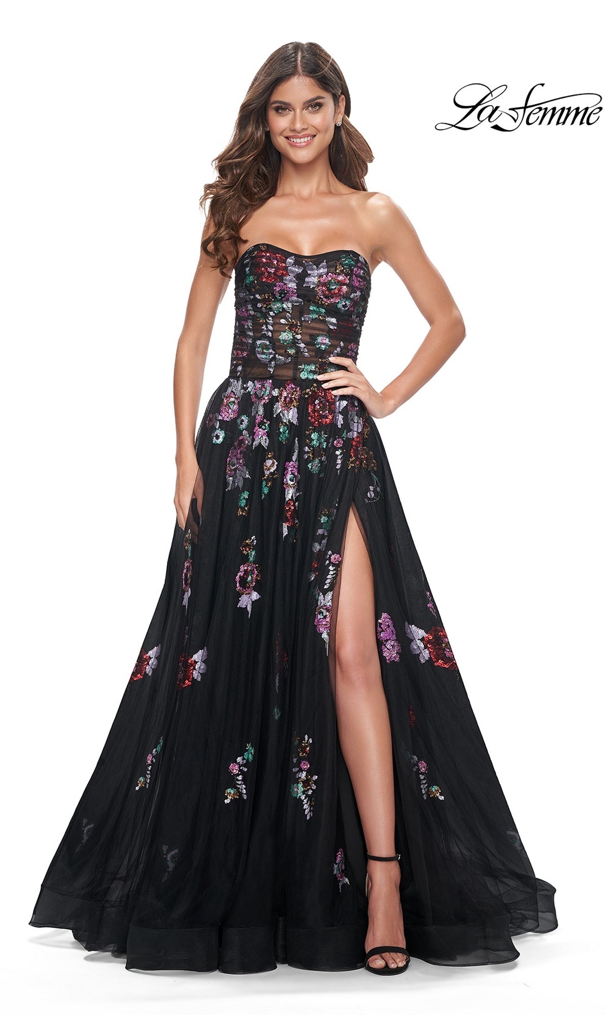 A woman wears the Le Femme Sequin-Floral Long Black Prom Dress 32072, featuring a strapless design, sheer bodice, floral embroidery, and thigh-high slit, standing against a white background with the