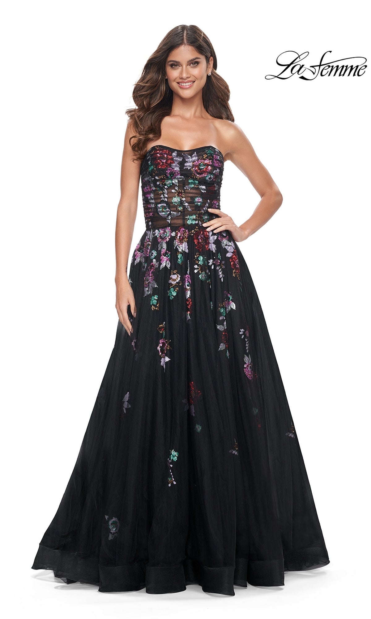 A woman stands against a white background wearing the Le Femme Sequin-Floral Long Black Prom Dress 32072 featuring a strapless sheer bodice with floral embroidery and a full tulle skirt. The