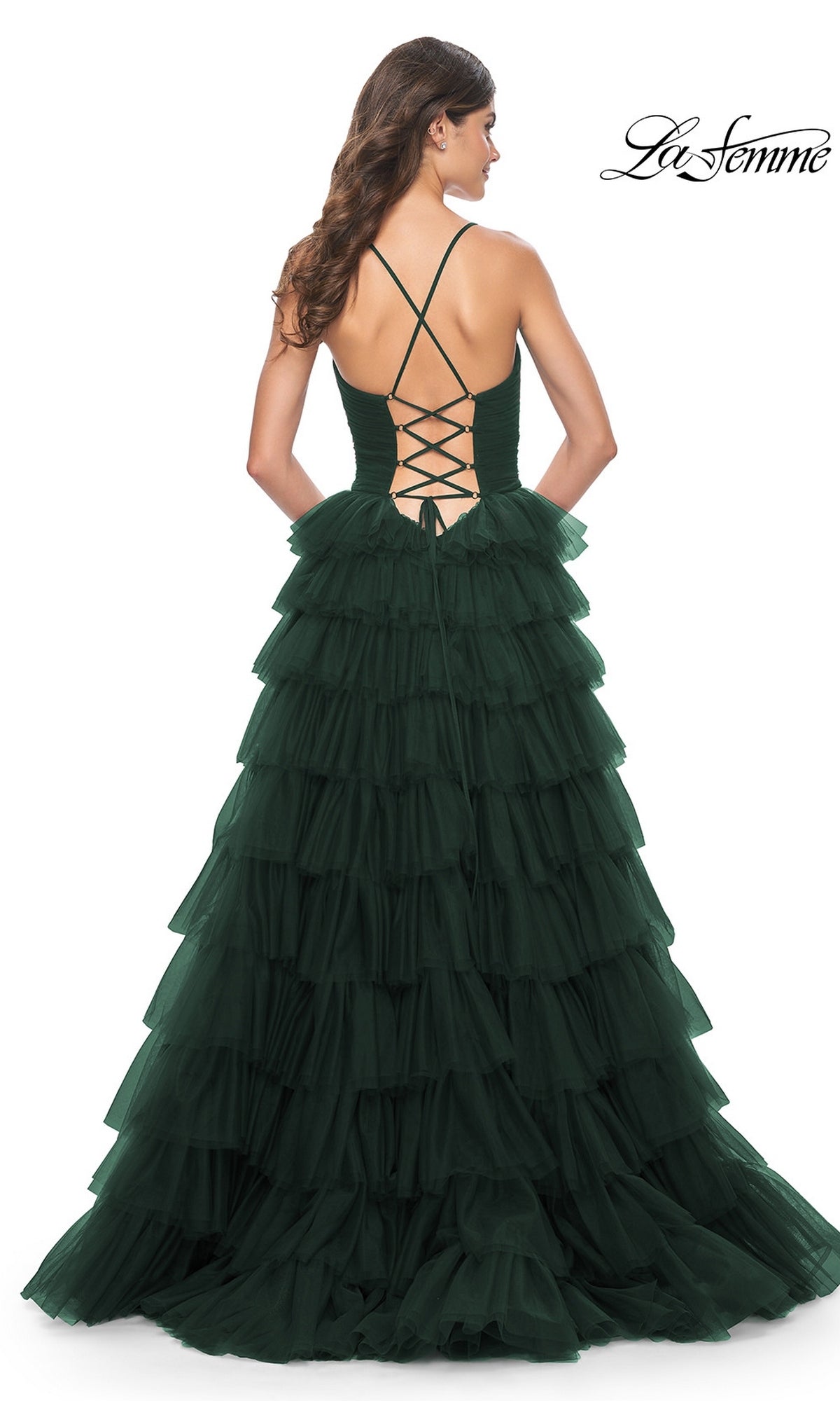 A woman wearing the La Femme Lace-Up Long Tiered Prom Dress 32086 in dark green stands facing away, highlighting the full length, ruffled tiers, and lace-up open back design of the gown.