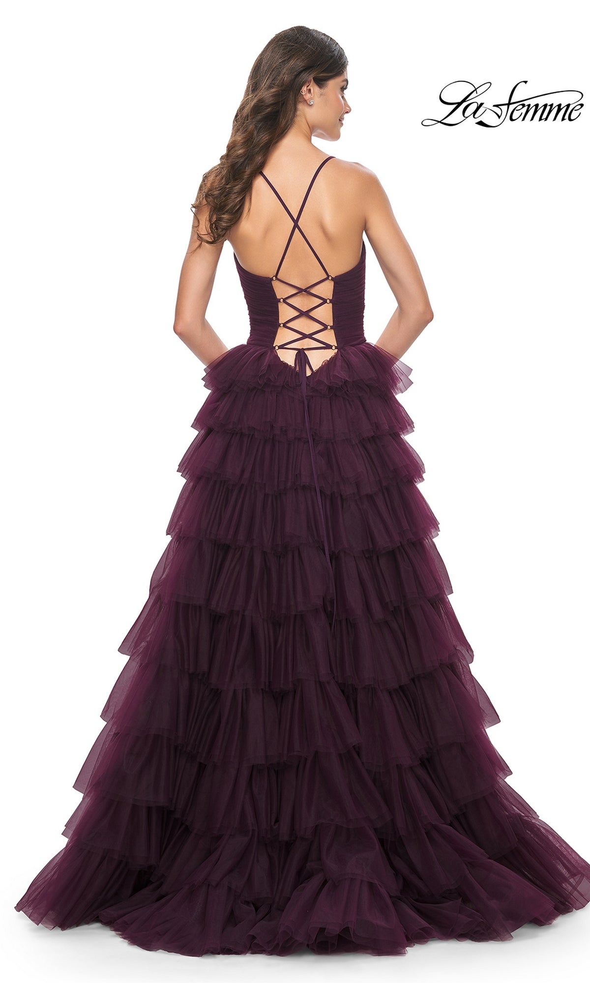 A woman wearing the La Femme Lace-Up Long Tiered Prom Dress 32086, a dark purple tulle gown with dramatic cascading tiers and an open lace-up back, stands facing away from the camera.