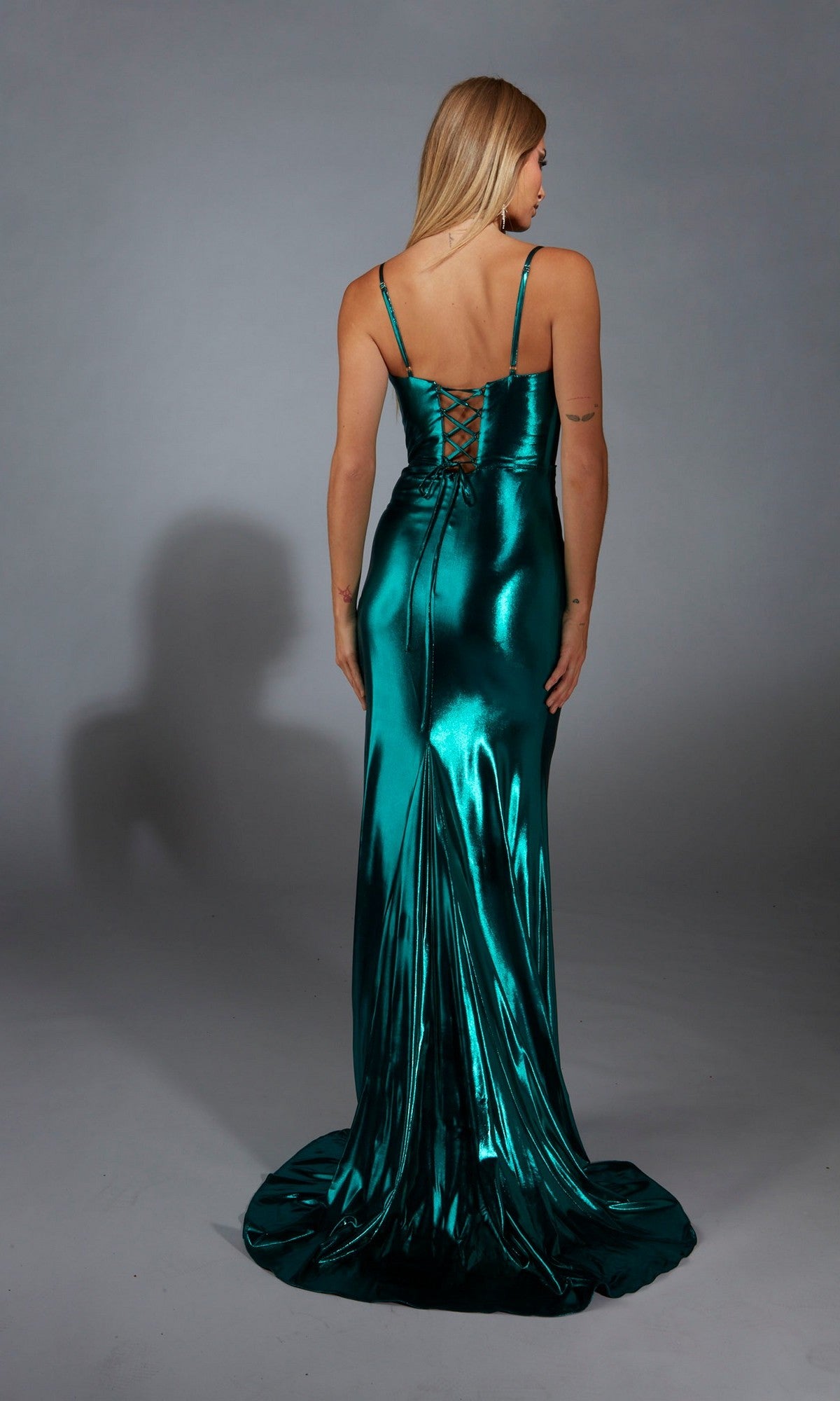 A woman in the Aerie Couture 320 long prom dress, featuring metallic green fabric, a cowl neckline, and a corset lace-up back, stands facing away against a plain gray background.