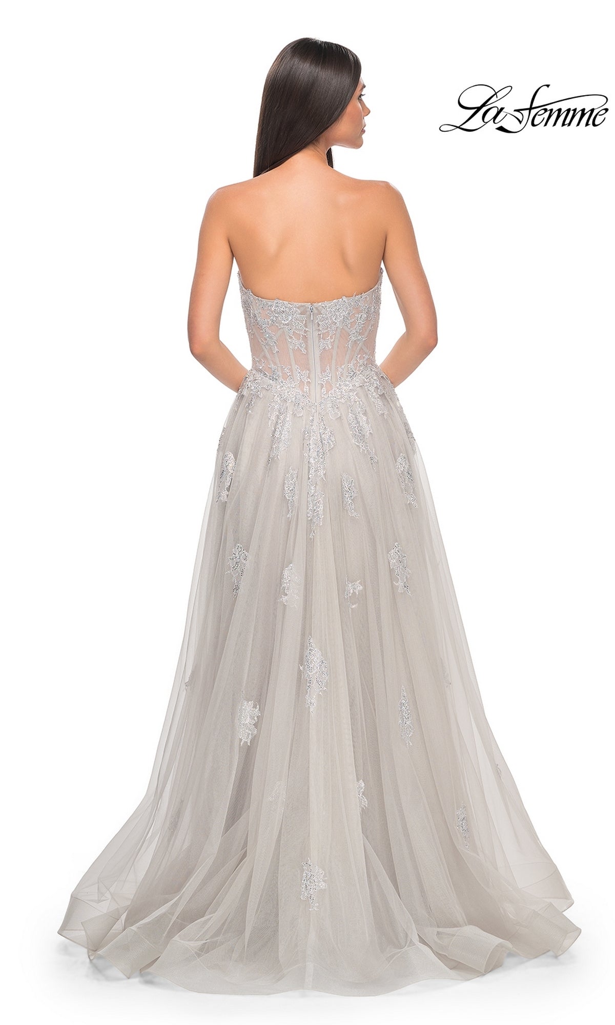 A woman stands facing away in the La Femme Strapless Long A-Line Prom Dress 32111, featuring silver embroidered-lace appliques, a sheer back, visible boning, and a flowing tulle skirt.