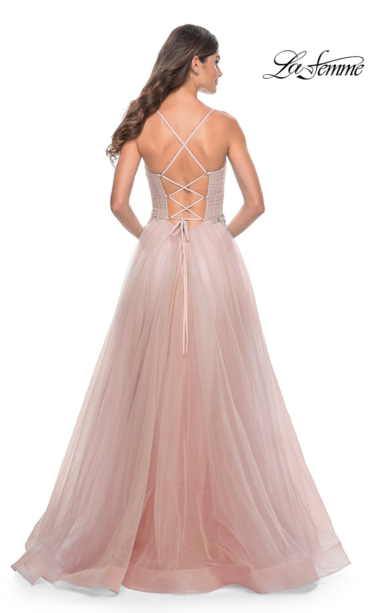 A woman stands facing away in the La Femme Sheer-Bodice Long A-Line Prom Dress 32117, featuring blush pink fabric, a lace-up corset back, and crisscross straps.
