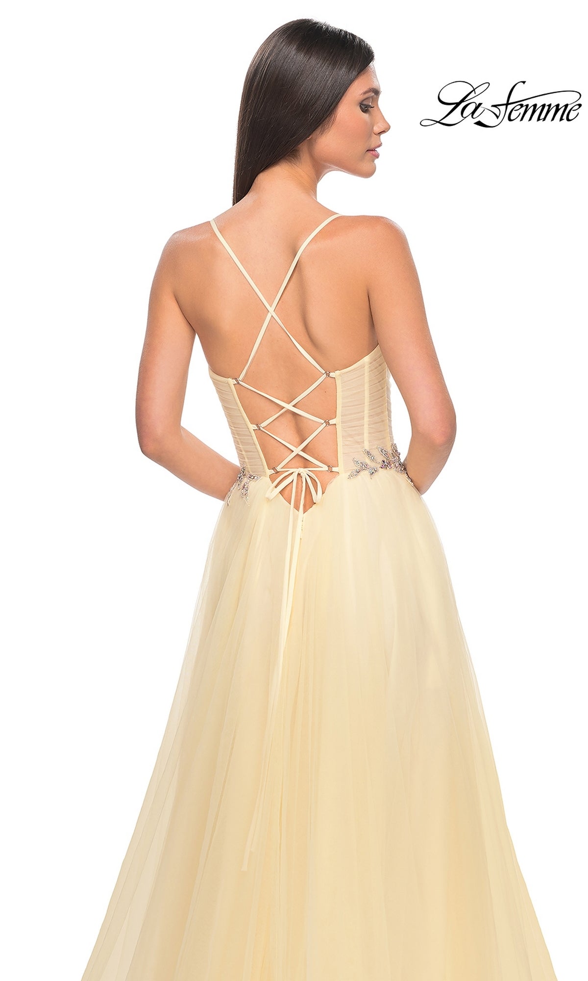 Woman wearing the La Femme Sheer-Bodice Long A-Line Prom Dress 32117 in light yellow, featuring a lace-up open back with thin crisscross straps and a flowing tulle skirt, standing against a white background.