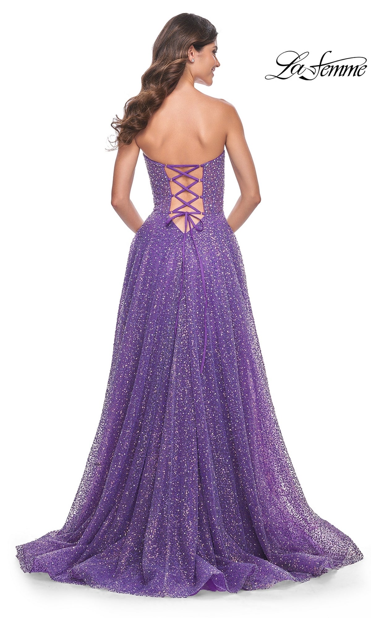 A woman stands facing away, wearing the La Femme Strapless Embellished Prom Dress 32136 in purple glitter, featuring an A-line silhouette, hot-stone embellishments, and a lace-up back.